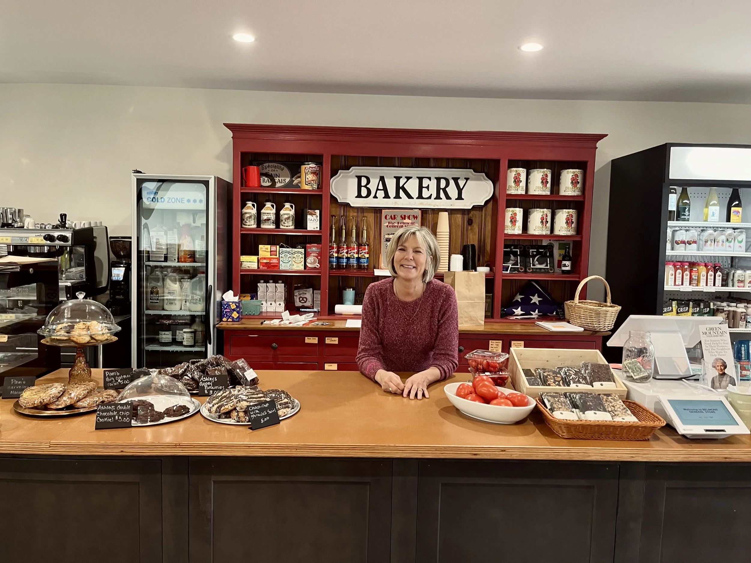 Smiling woman behind bakery counter with various baked goods and tomatoes, bakery sign, and shelves with cans and products.