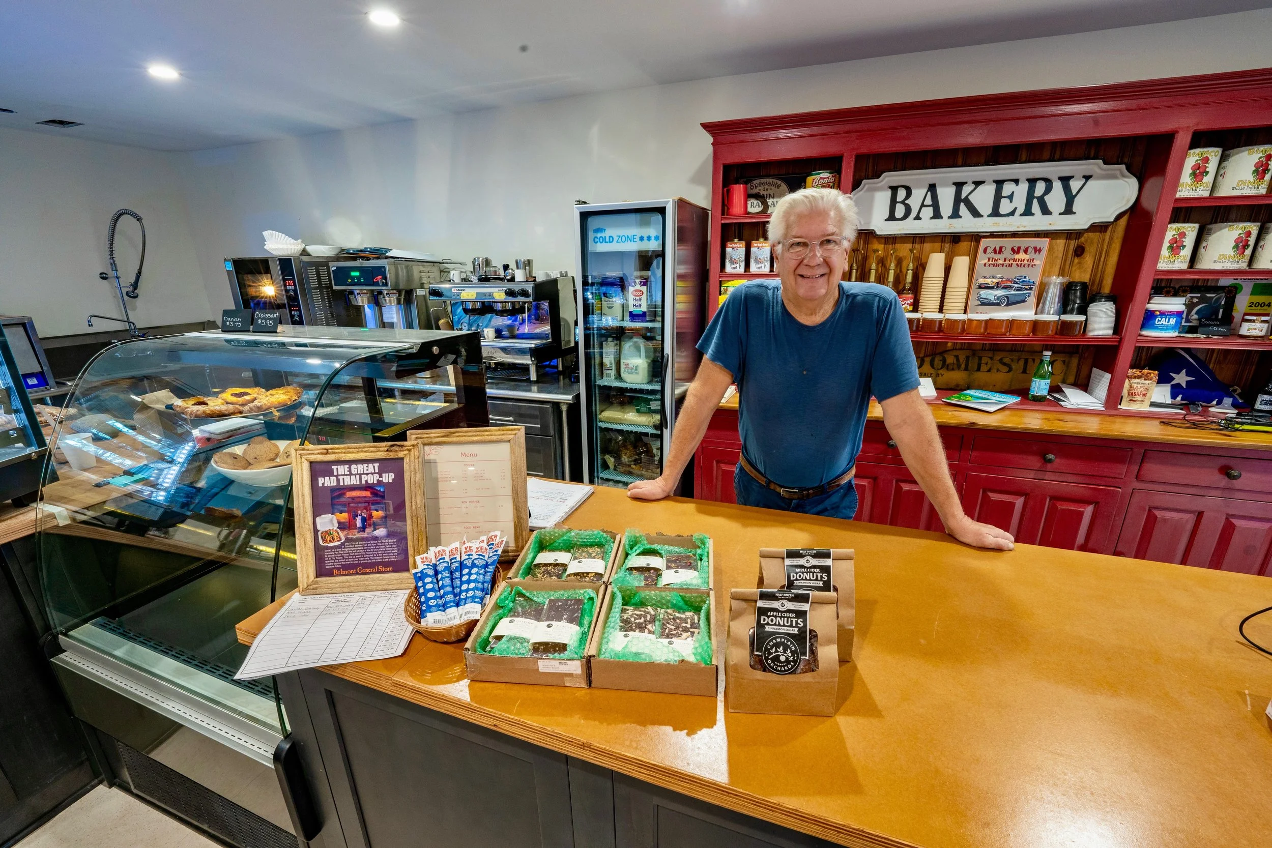 A smiling elderly man behind a bakery counter with baked goods, in a store with bakery signs and shelves of products.
