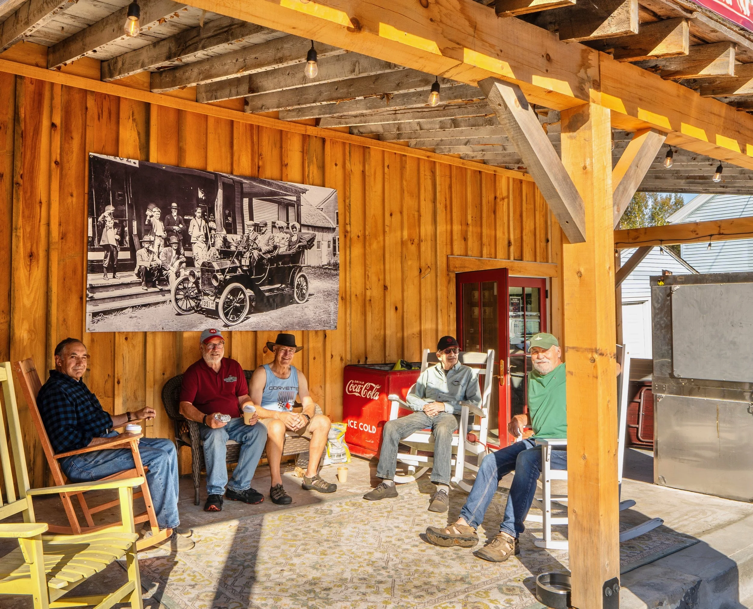 Five men sitting on chairs and a bench under a wooden patio, with a large black-and-white historical photograph on the wall behind them. The men are holding drinks, and the patio has a rug and a Coca-Cola cooler. The setting appears to be outdoor and