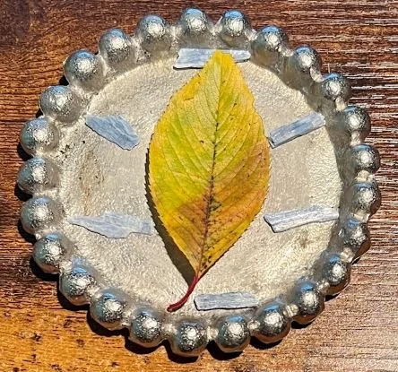 A grid with healing energy, with a yellow leaf in the center on a silver circle. There are six small pieces of blue kyanite crystal surrounding the leaf