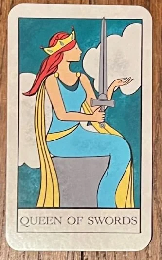 The Queen of Swords Tarot Card, a woman with red hair sitting on a concrete stone holding a sword in her right hand
