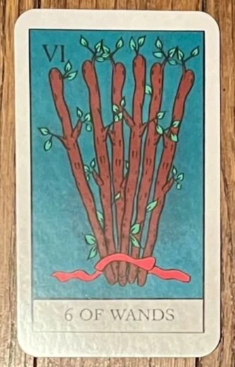 The six of wands tarot card with six wooden wands from a tree bundled together with a red ribbon at the base