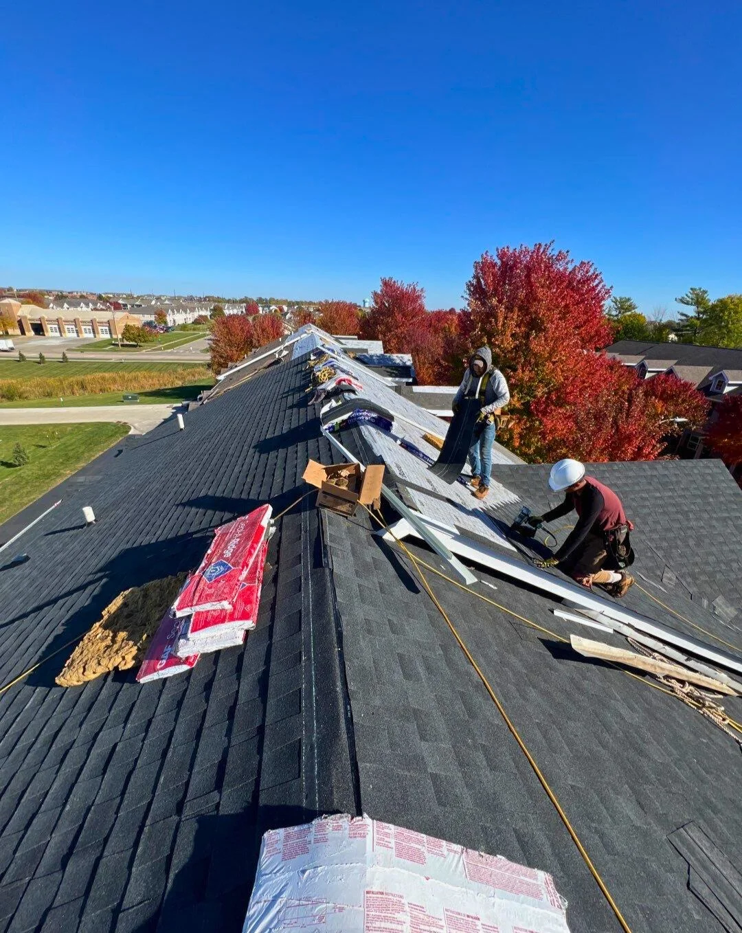 Roof workers repairing shingles on a sloped roof with autumn trees and a neighborhood in the background.