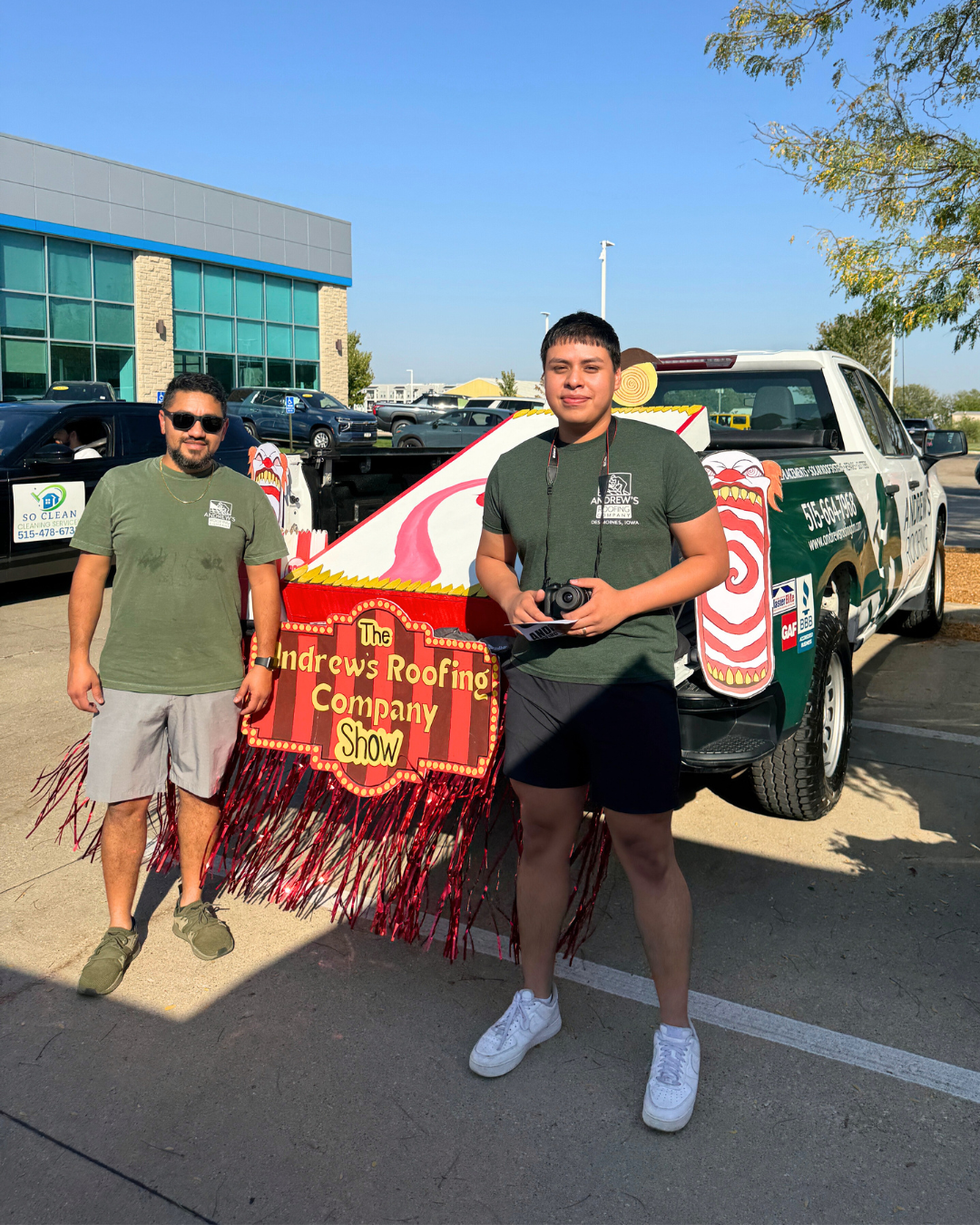 Andrew's Roofing Waukee - Two men standing in front of a pickup truck with a sign for The Andrew's Roofing Company Show, with a carnival game inside a colorful booth, in a parking lot on a sunny day.