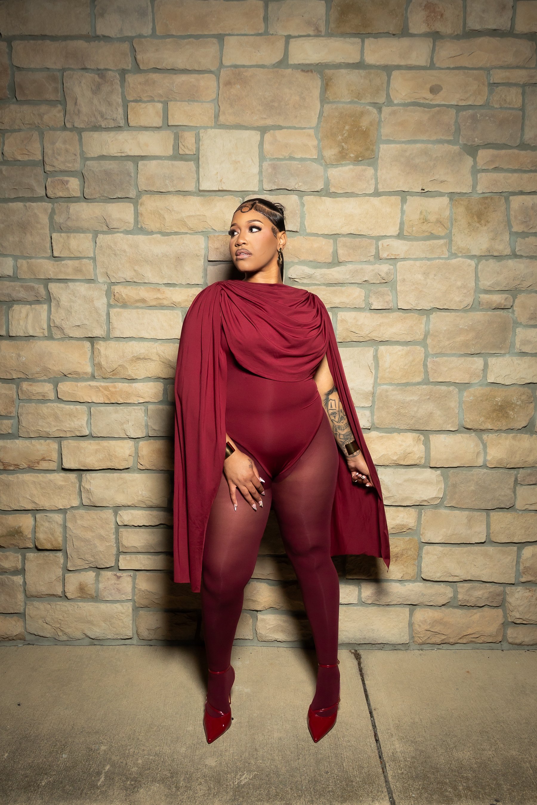 A woman dressed in a burgundy sheer bodysuit, matching high heels, and an elegant cape poses against a stone wall.