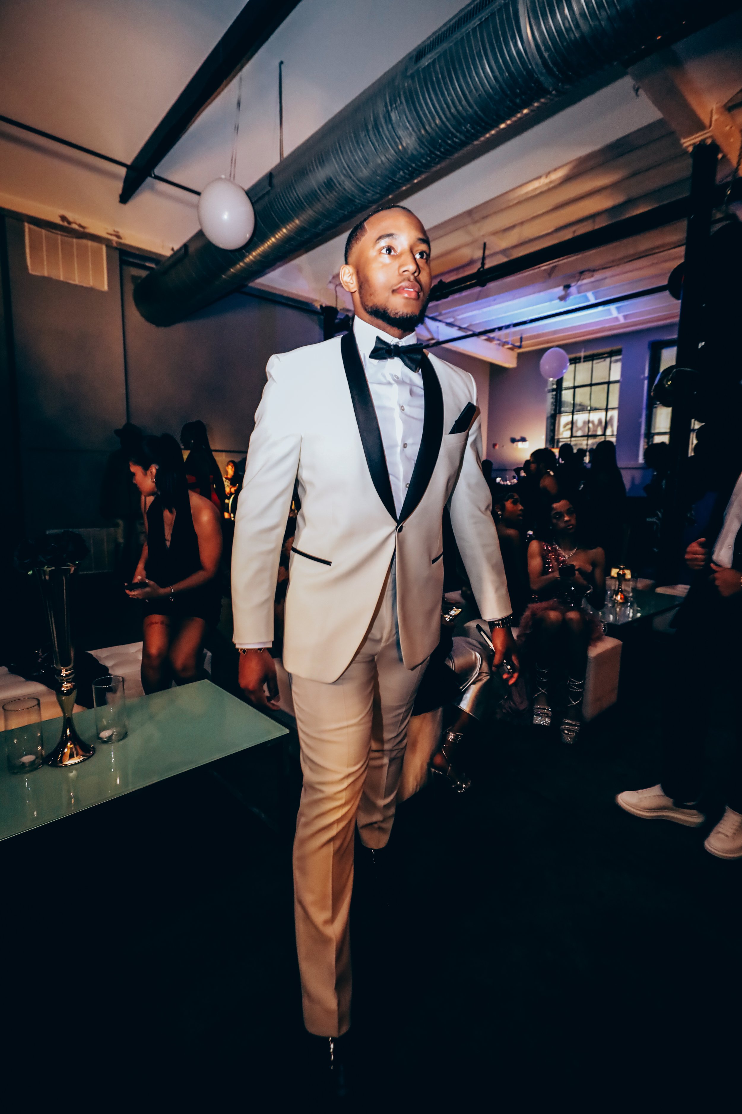 A man in a white tuxedo with black lapel and bow tie stands in a dimly lit party or club, surrounded by other guests sitting on couches and standing nearby, with modern decor and large industrial-style windows in the background.