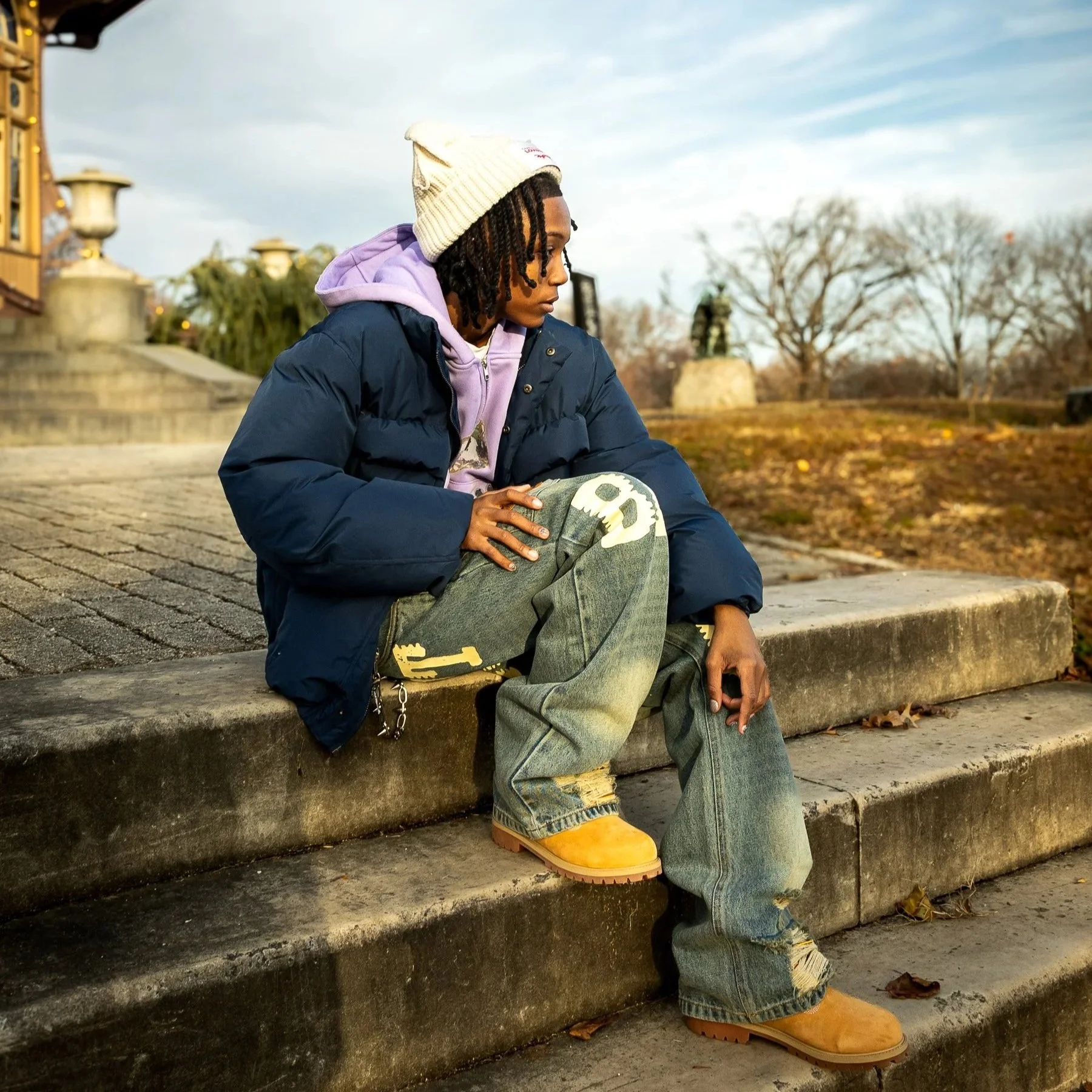 A young man with dreadlocks sits on concrete steps outdoors on a cloudy day, wearing a white beanie, purple hoodie, dark puffer jacket, distressed jeans, and tan work boots, with leafless trees in the background.