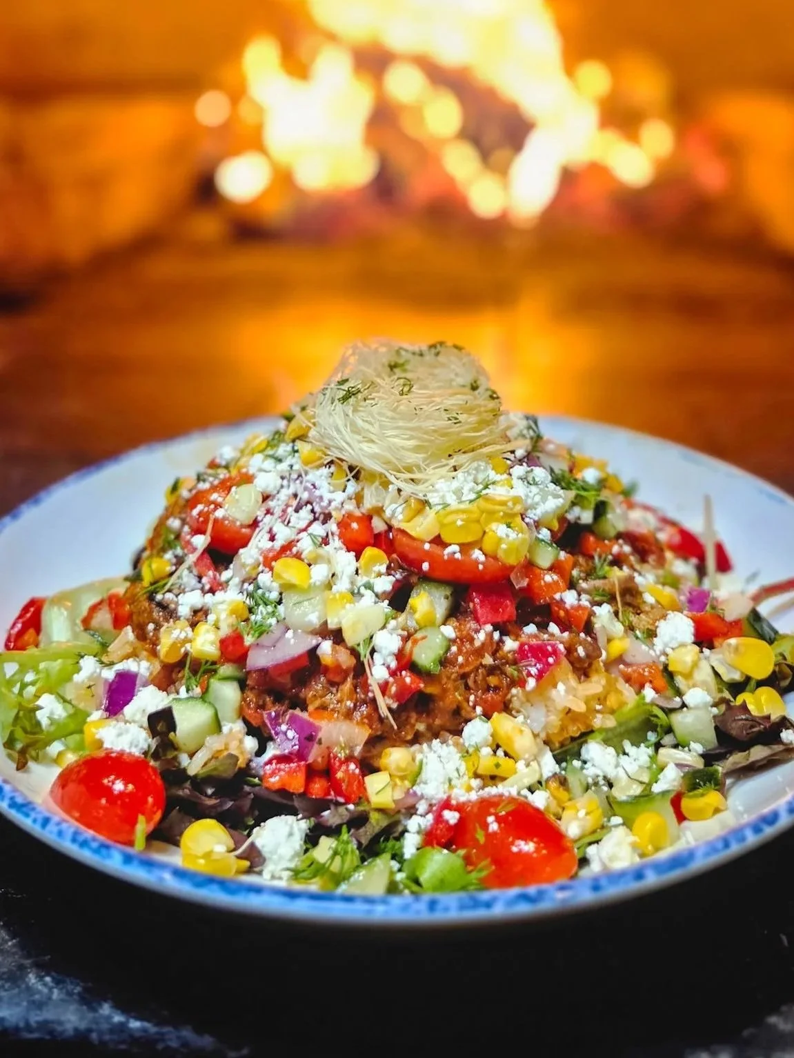 A colorful braised lamb bowl with fresh vegetables, corn, cheese, and shredded lamb, topped with kadayif and herbs, served in a blue-rimmed white bowl on a dark surface with a blurred fire in the background.