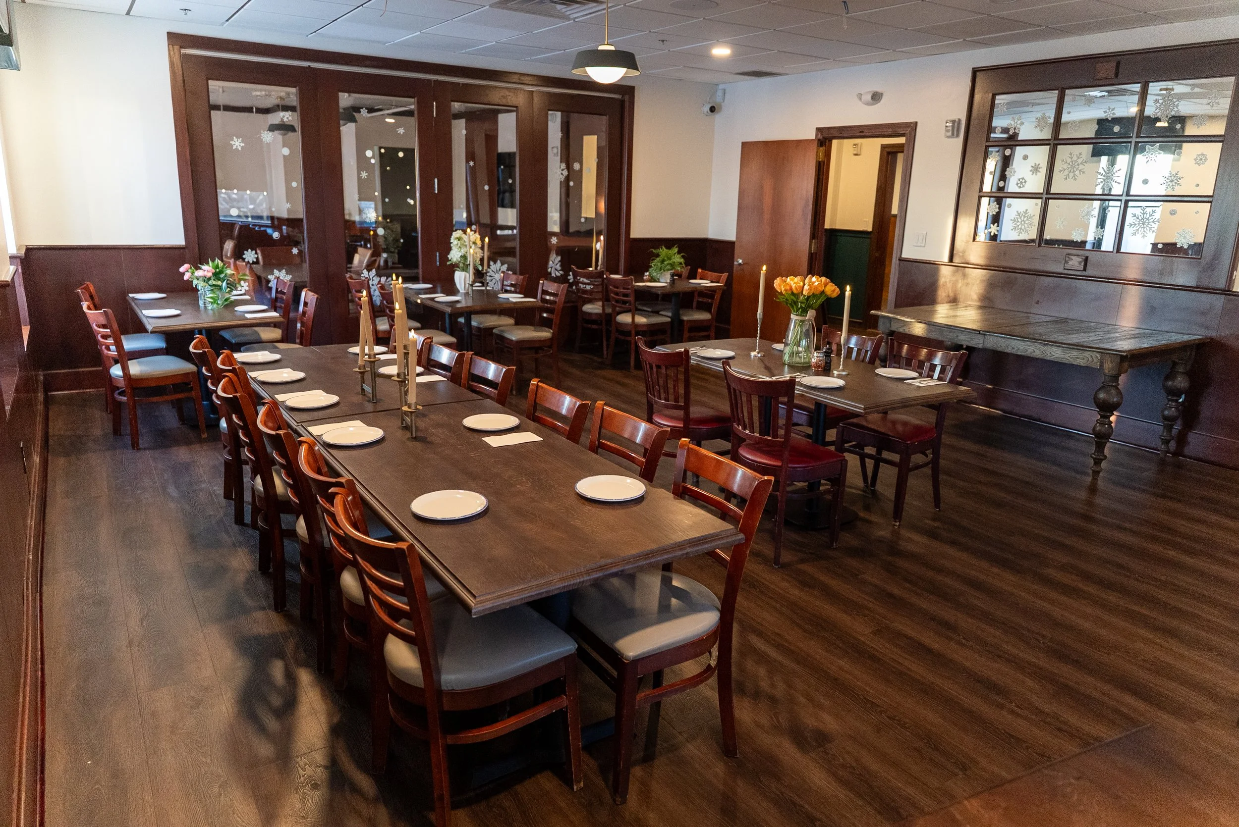 A restaurant dining area with wooden tables and chairs, set with white plates and napkins, decorated with flowers and candles, and snowflake decals on the windows.