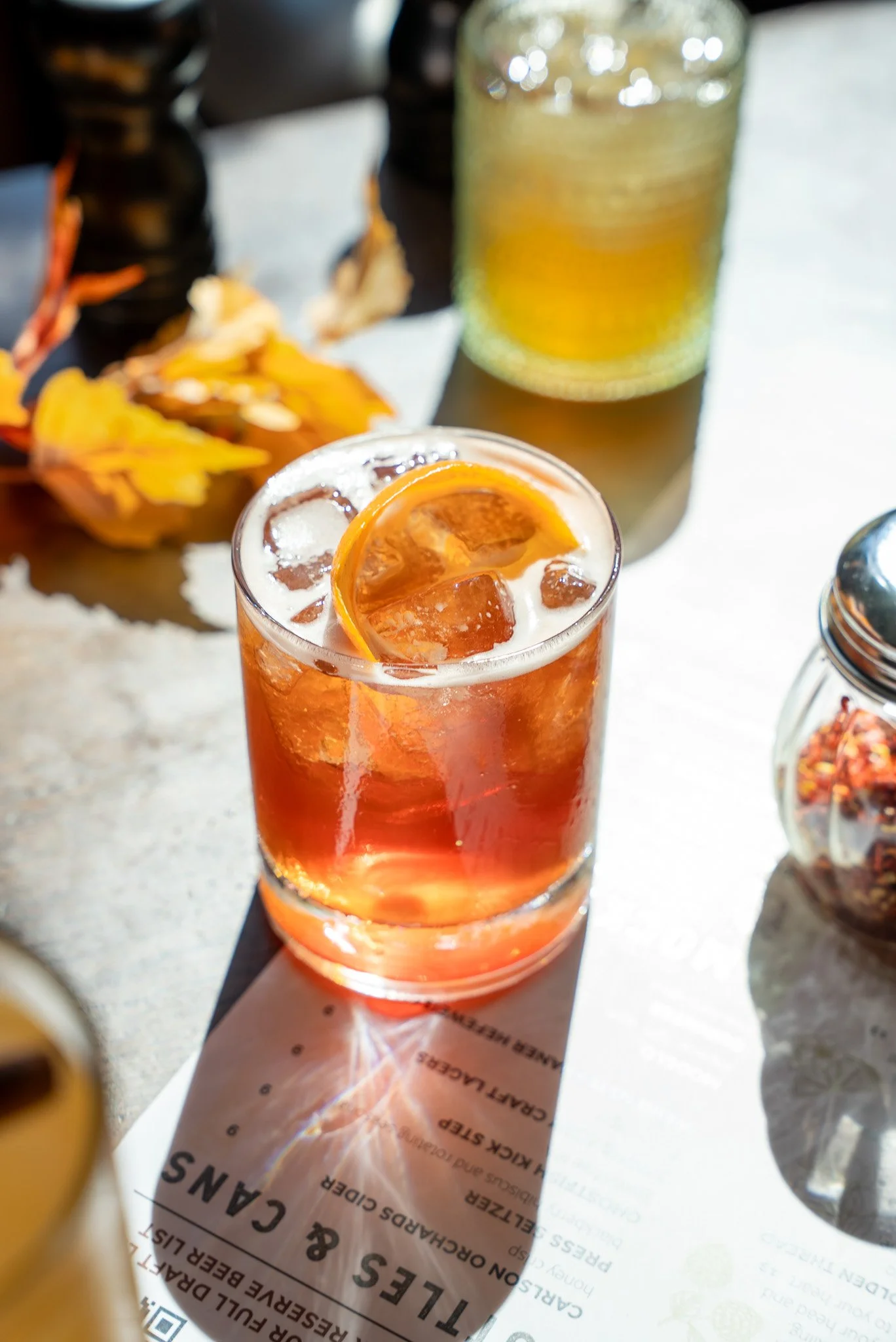 A glass of iced tea garnished with an orange slice sits on a table next to a menu with a blurred background of a beer bottle, a green bottle, and string lights.