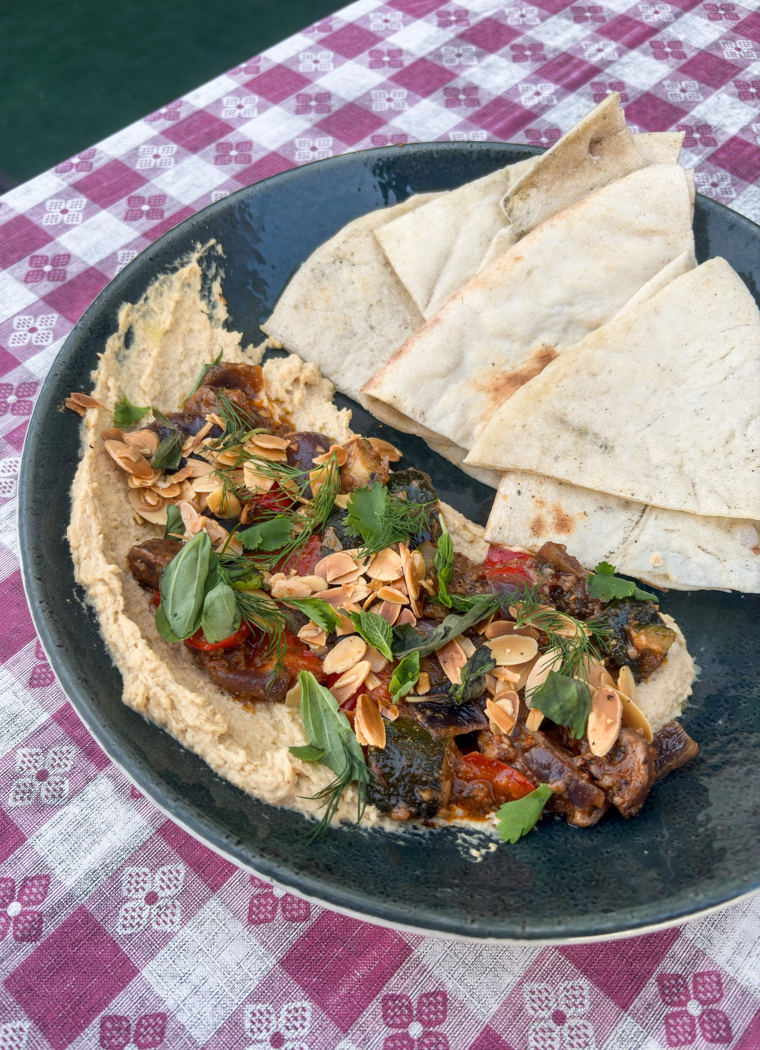 A black plate of lemon & herb hummus, pita bread, and garnished with herbs and sliced almonds, on a red and white checkered tablecloth.