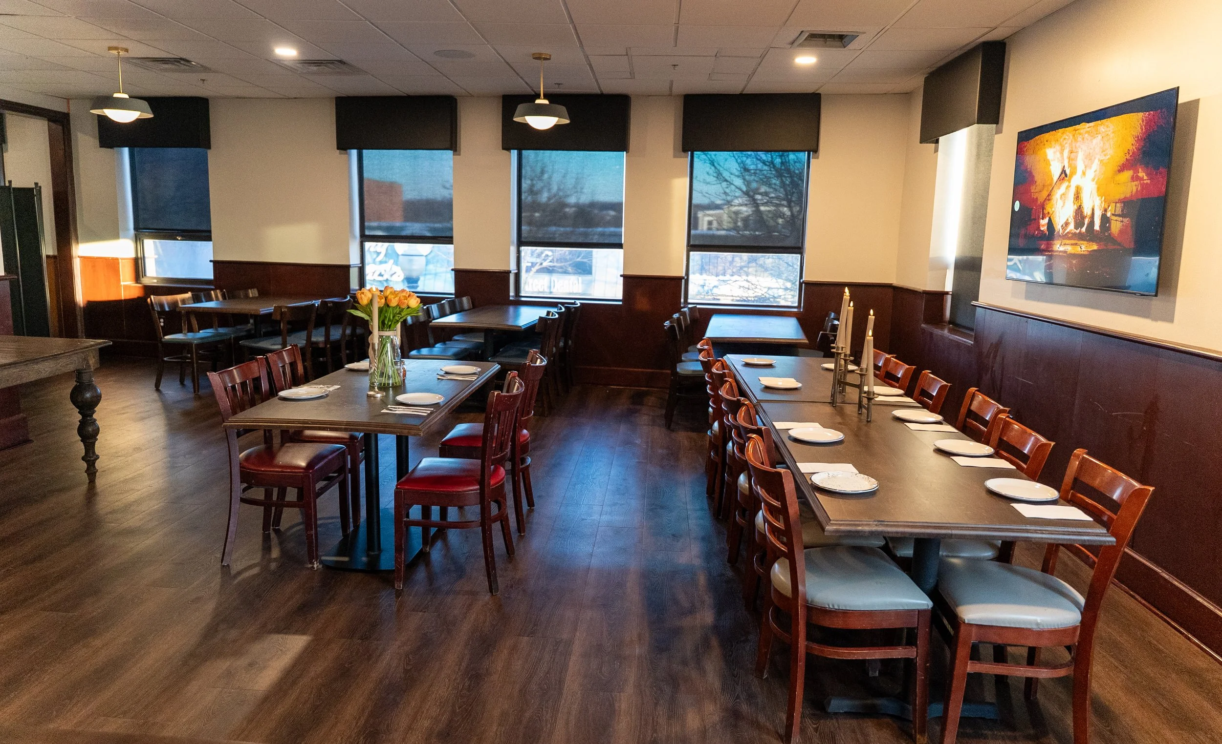 Empty dining room with dark wooden floors, a long main table set with white plates and napkins, surrounded by wood chairs with padded seats, and a smaller table with a vase of yellow and orange tulips. Windows with black shades, a flat-screen TV on t