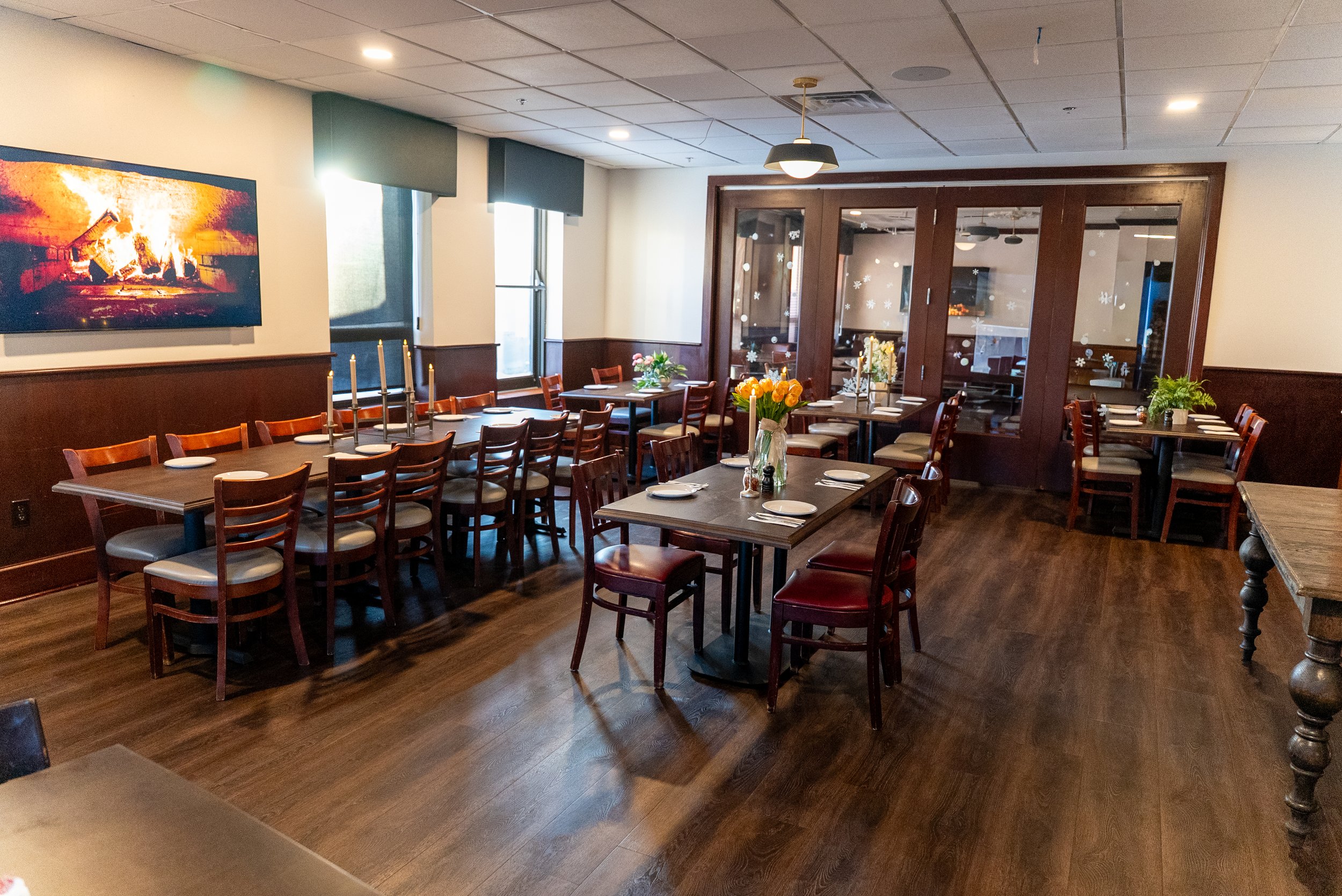 A cozy restaurant dining area with wooden tables and chairs, decorated with flowers, candles, and place settings, with large windows and a TV on the wall.