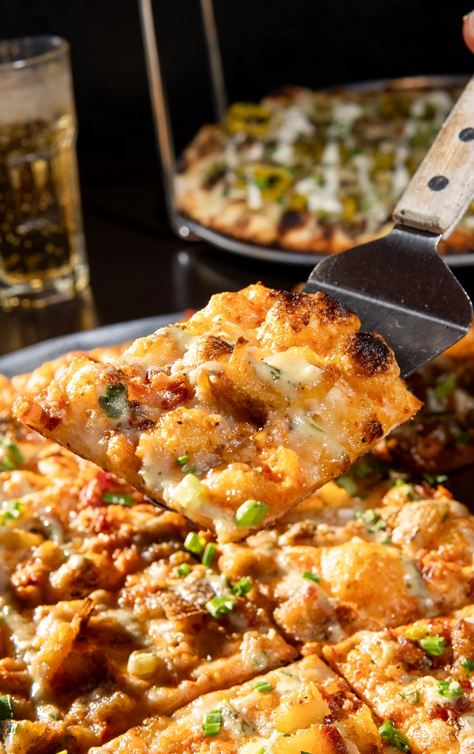 Close-up of a slice of pizza with cheese, sausage, and green onions, being lifted from a hot pizza tray. In the background, a pizza and a glass of soda are visible.