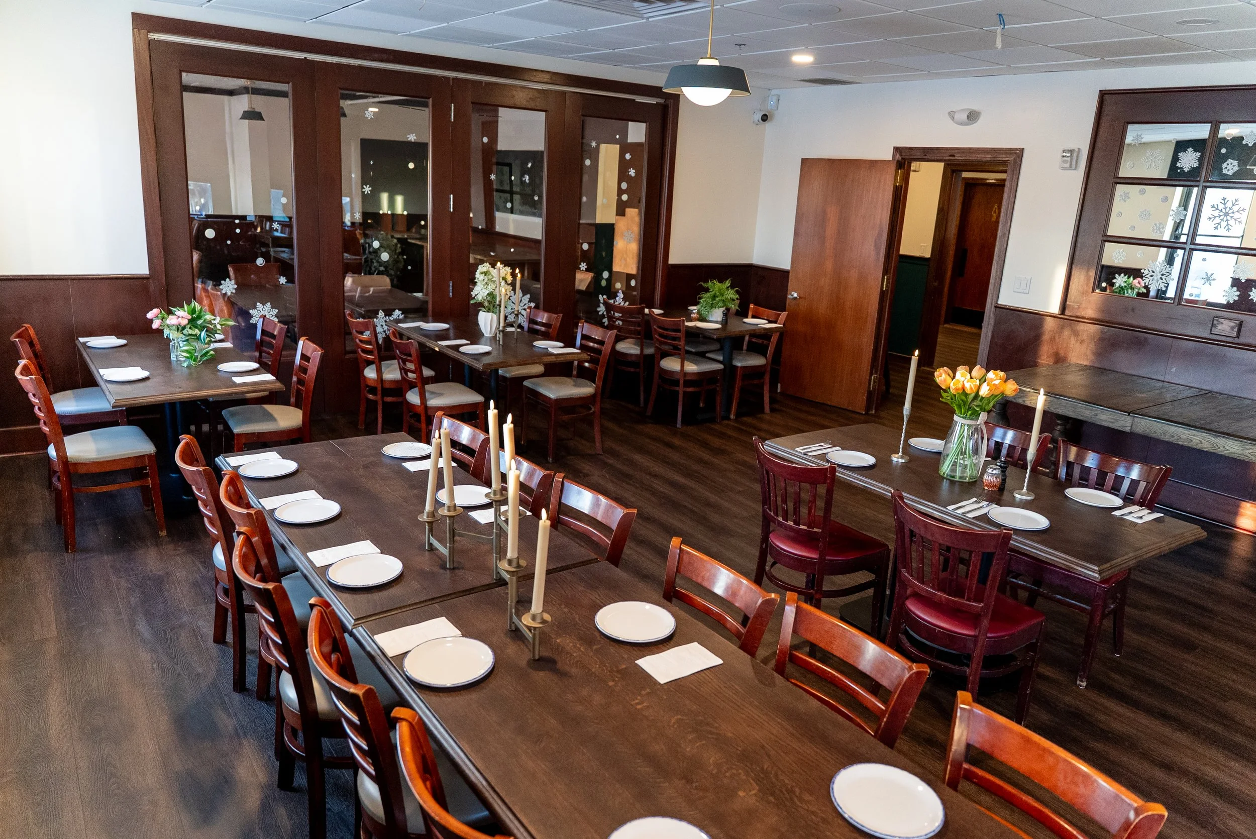 A cozy dining room decorated for a celebration with long wooden tables set with white plates, napkins, and silverware, adorned with floral centerpieces and tall candles. The room has dark wood flooring, paneled walls, and windows with snowflake stick