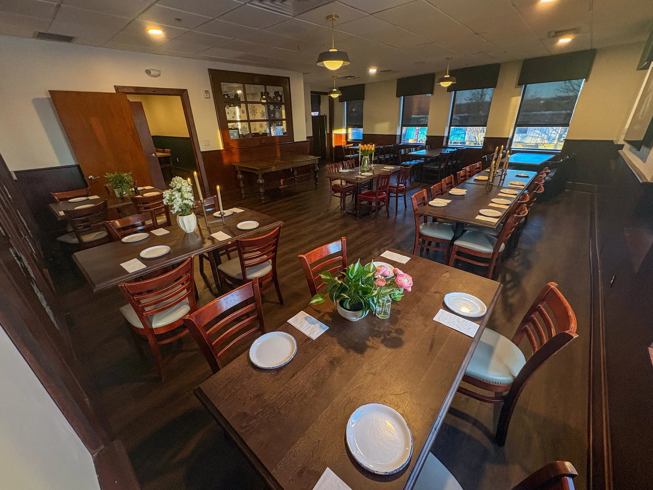 Interior of a cozy restaurant dining area with wooden tables set with plates, glasses, and flowers, surrounded by wooden chairs, with large windows and soft lighting.