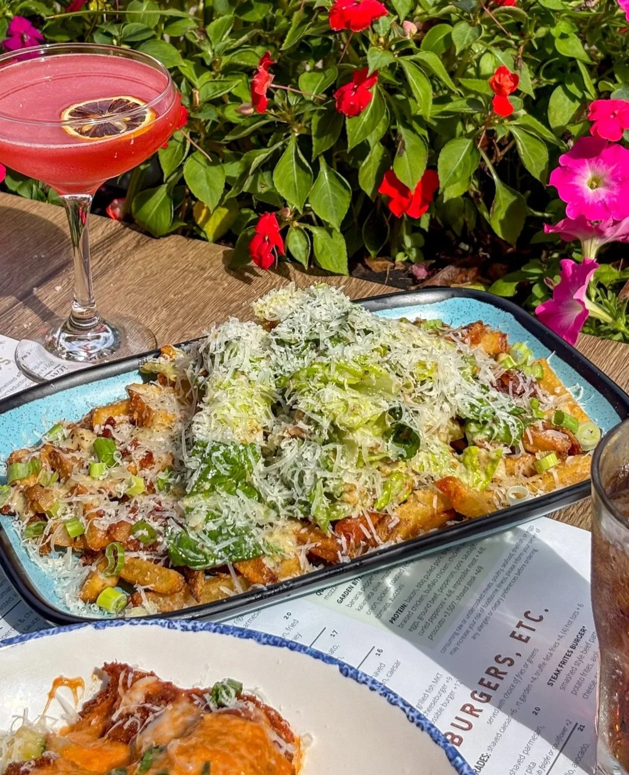 A plate of loaded fries topped with shredded cheese, chopped green onions, and shredded lettuce, with a pink cocktail garnished with lemon slices in the background and colorful flowers on a wooden table.