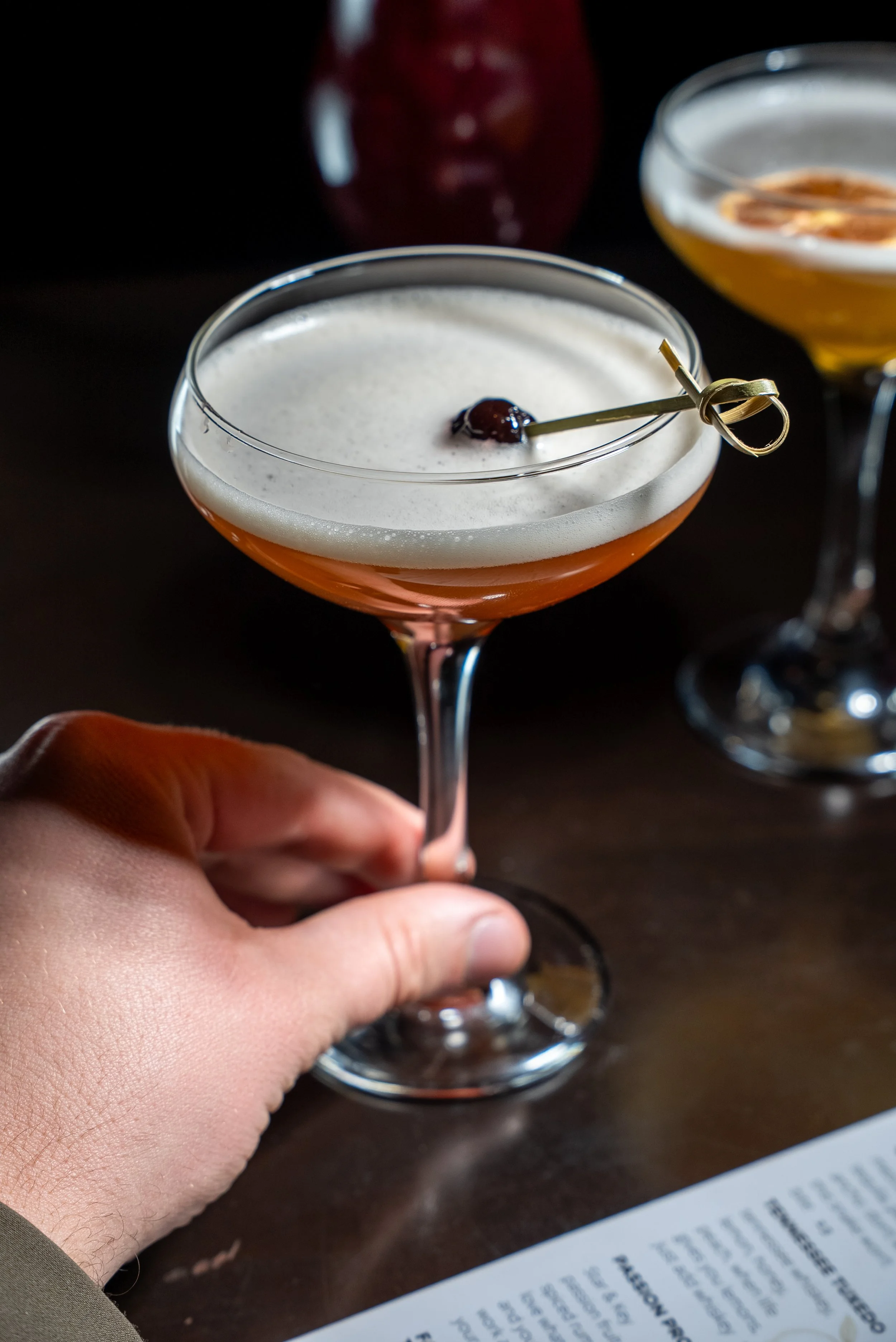 A hand holding a cocktail glass with an orange-colored drink topped with foam and garnished with a cherry on a toothpick. There is a blurred menu and another cocktail in the background.