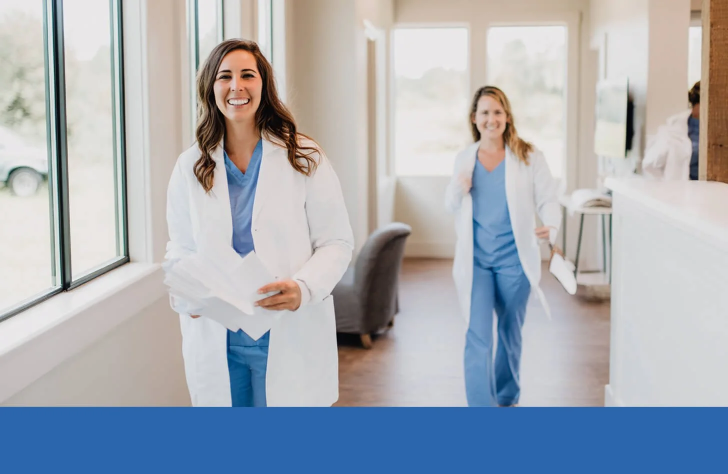 Doctor wearing a long-sleeved white lab coat over blue medical scrubs in a clinical healthcare setting, representing professional hygienic apparel for medical staff.