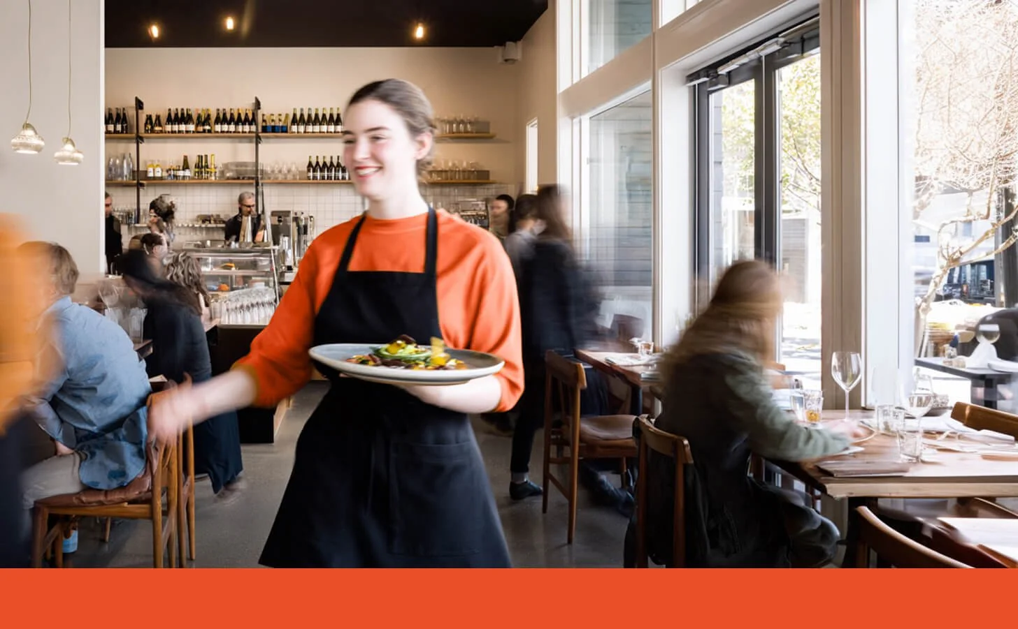 A Pacific Northwest restaurant server, wearing a black bib apron, serving plated food in a bustling restaurant.