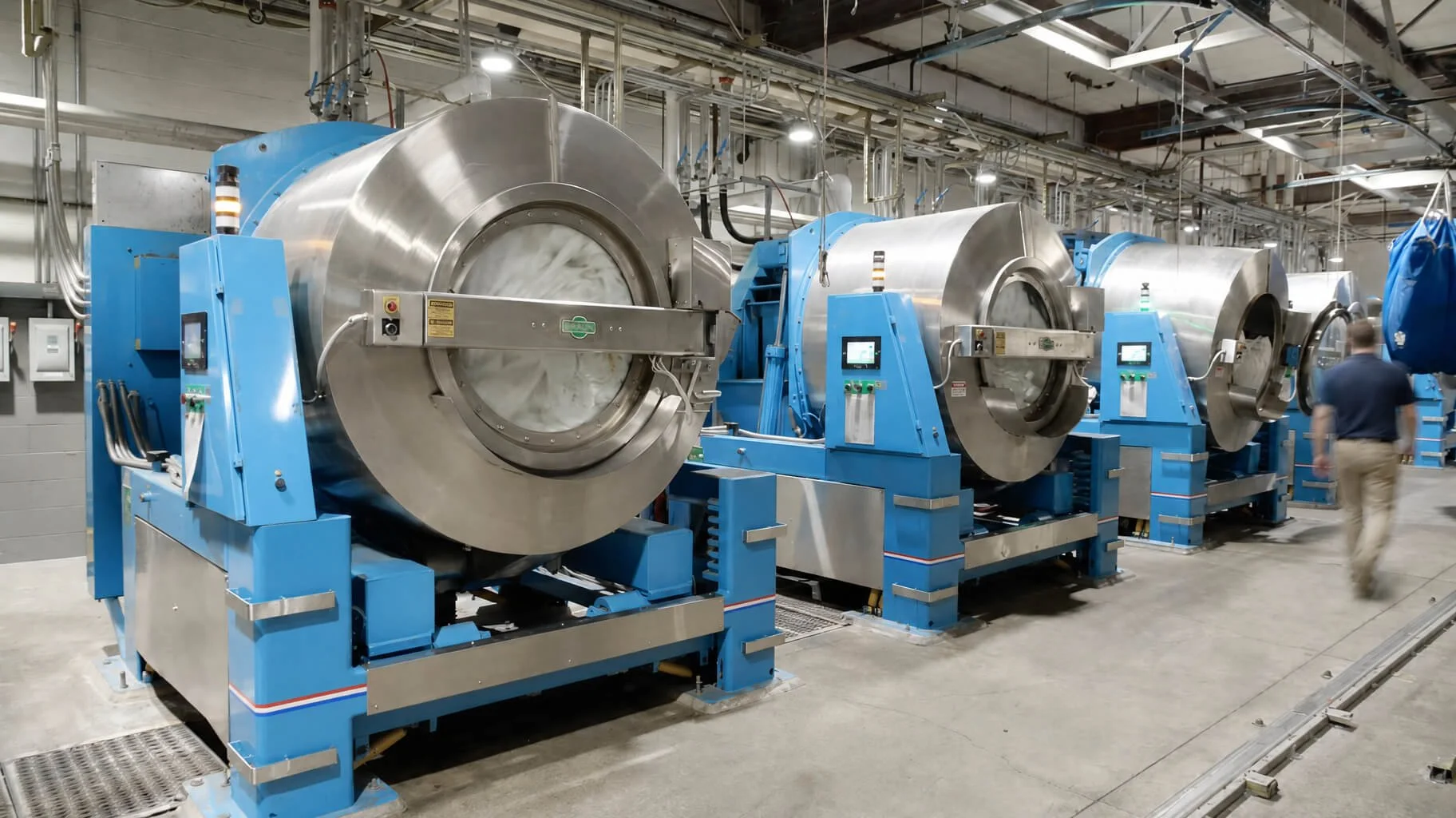 Industrial laundry facility with a row of large commercial washing machines and an employee walking through the production floor.