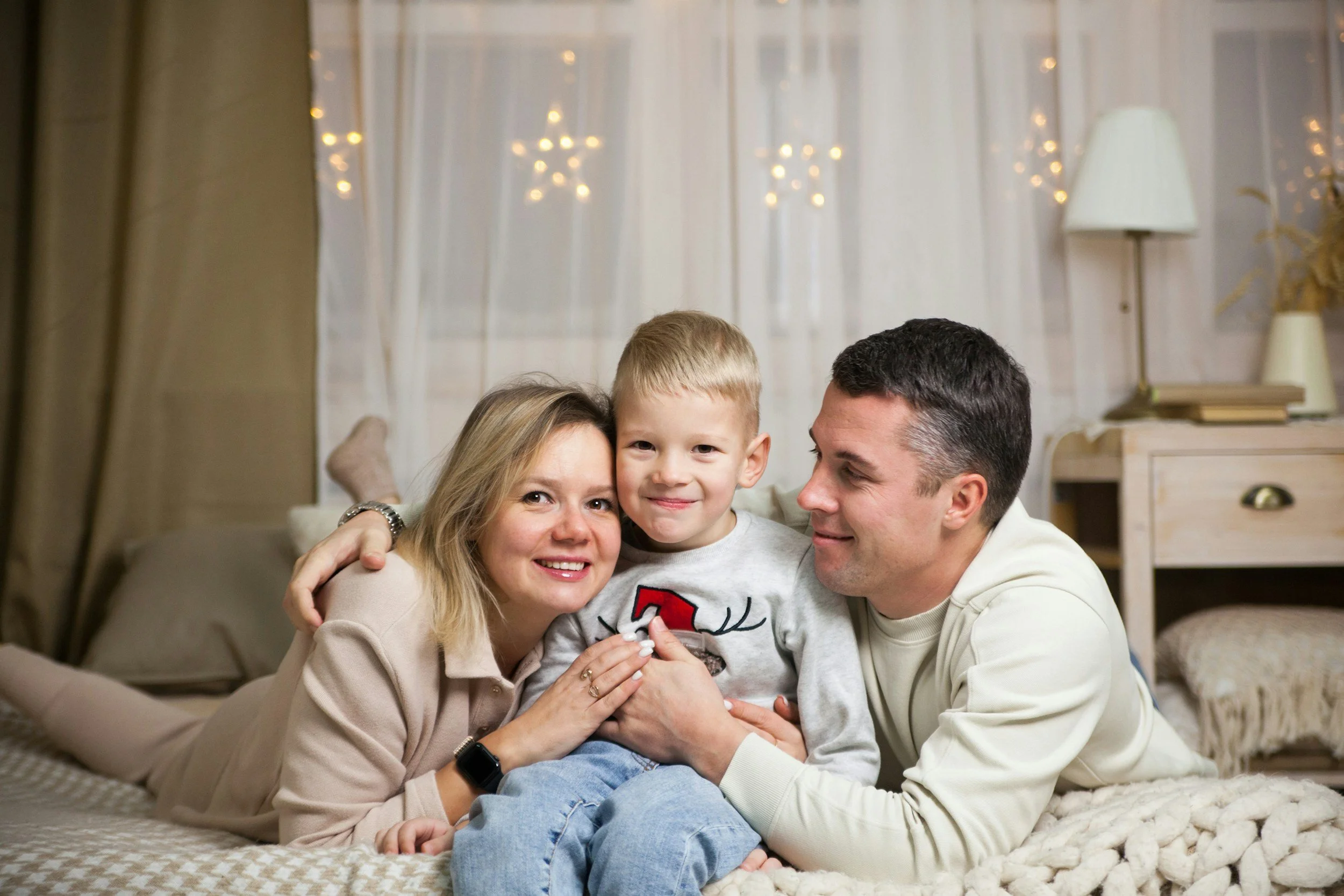 Family of four sitting on the floor in a cozy living room with white curtains and decorative star-shaped lights, smiling and enjoying each other's company.