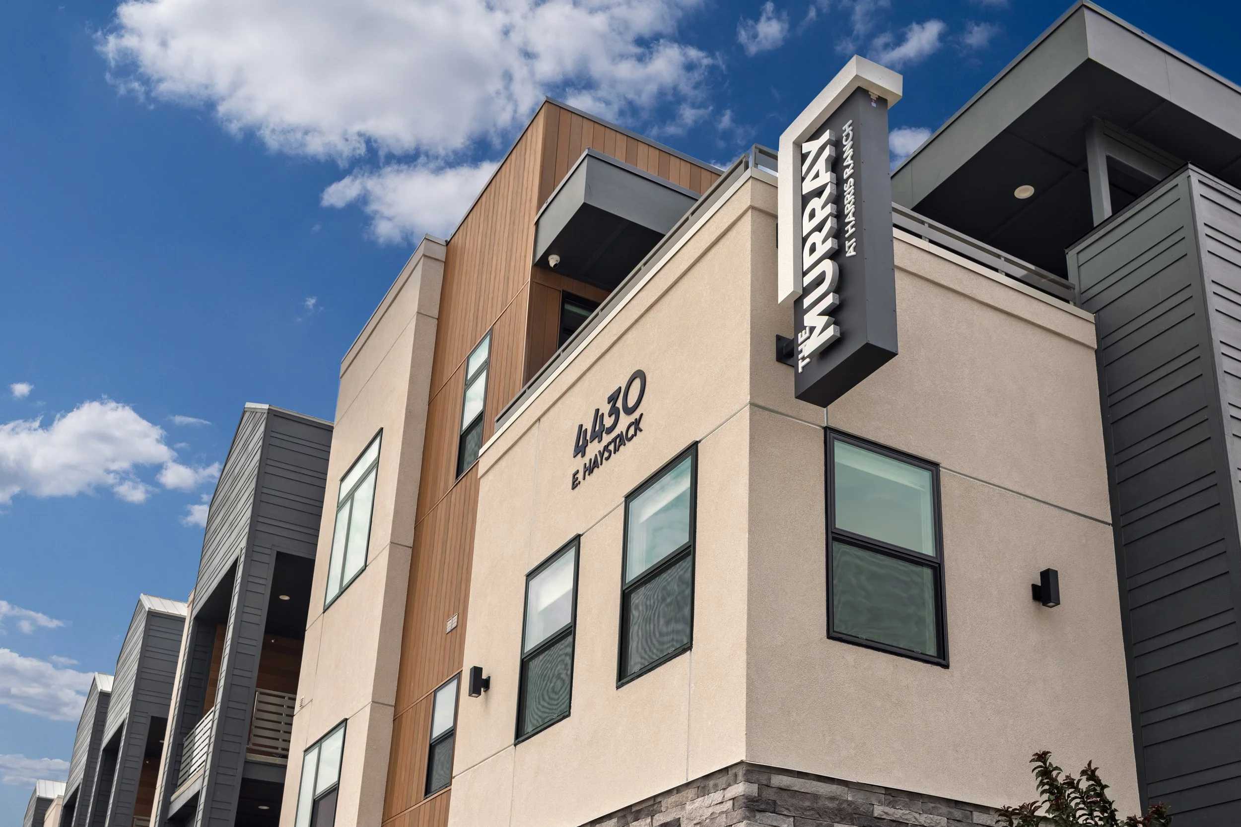 A modern multi-story apartment building with a sign reading 'THE WAVERLY E. HASTACK,' featuring beige, gray, and wood exterior with black framed windows, under a partly cloudy blue sky.