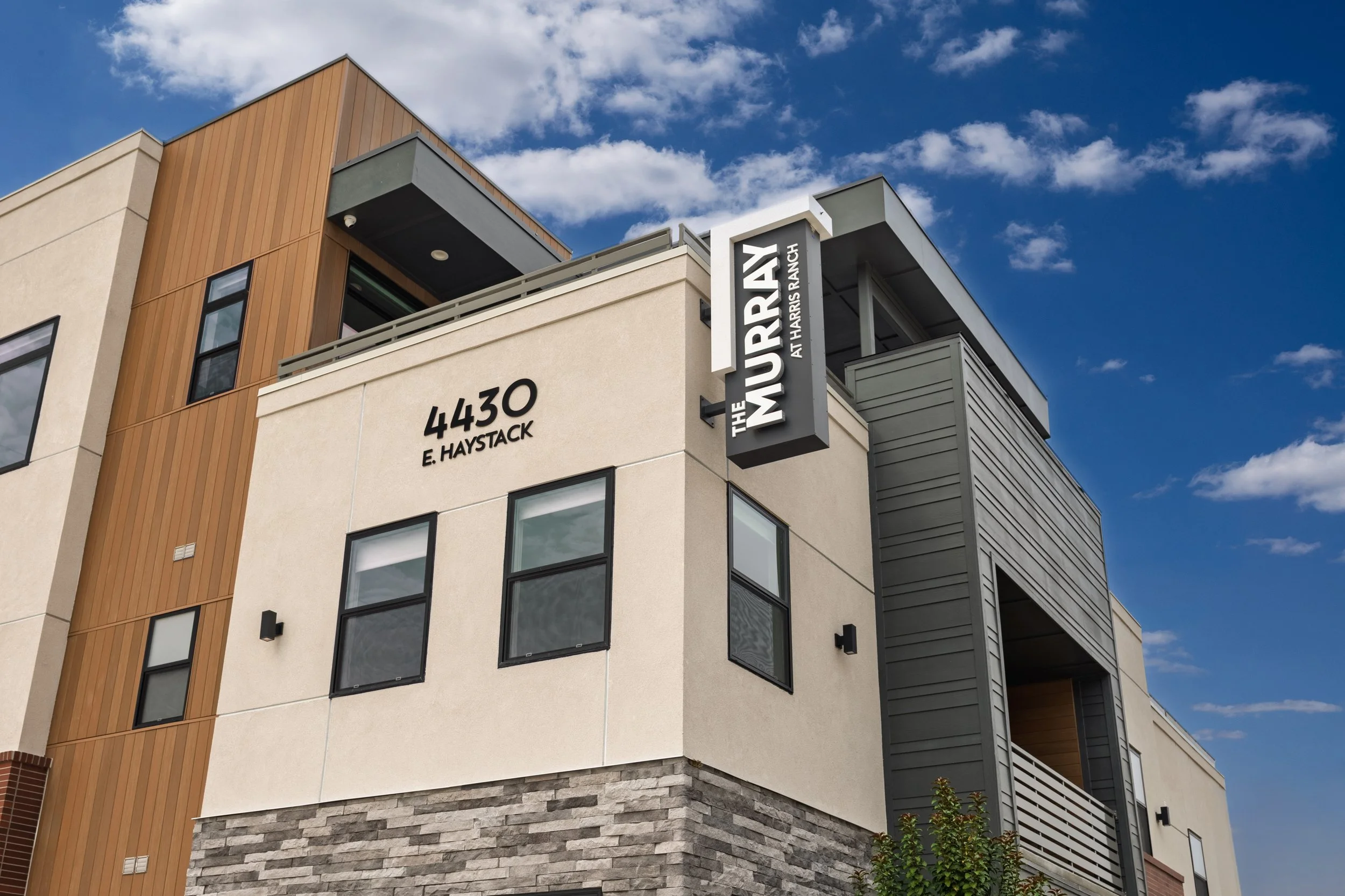 A modern, multi-story building with beige, gray, and brown exterior materials, with a sign that reads "The Murrray at Harpers Ranch" and the address 4430 E. Haystack, under a blue sky with some clouds.
