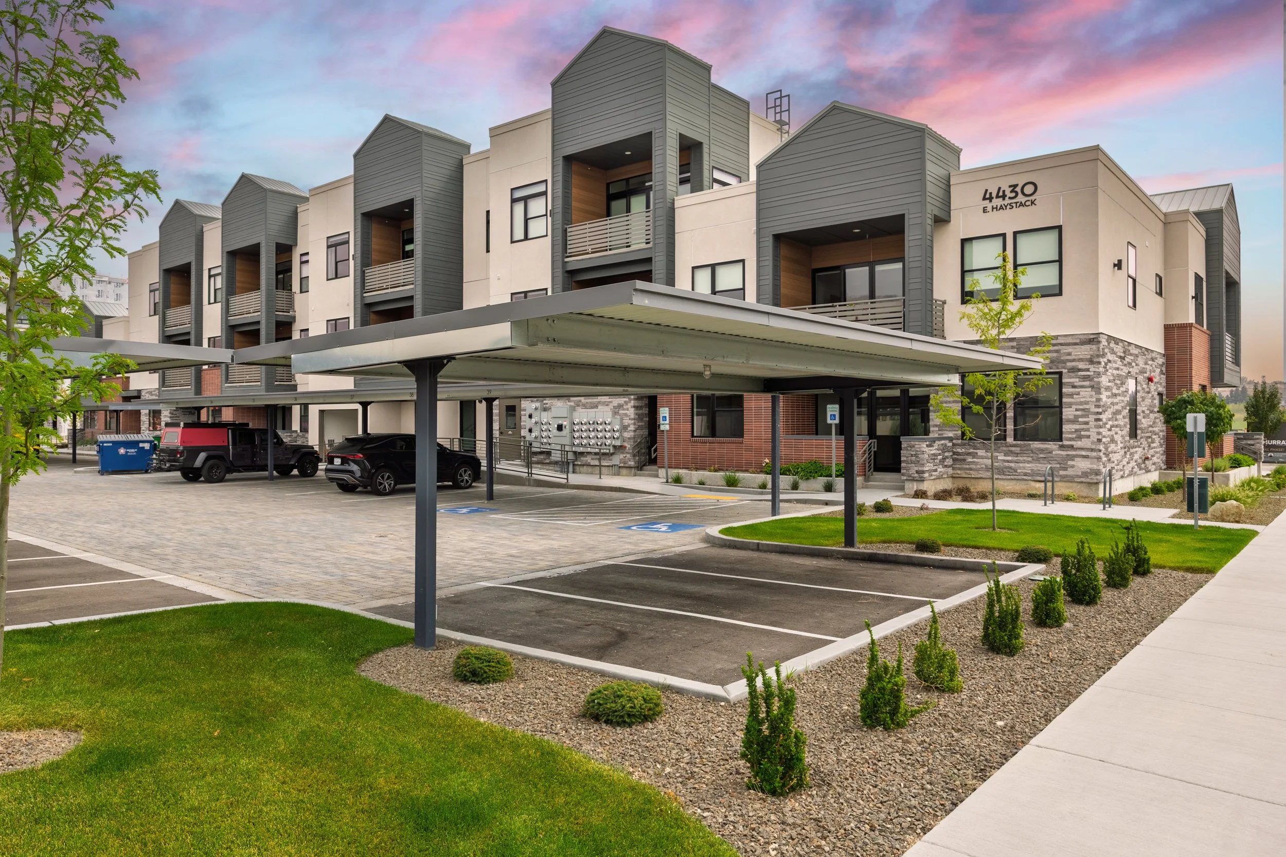 View of a modern multi-story apartment building with parking lot in front, covered parking structure, green lawn, and trees at sunset with pink and purple sky.