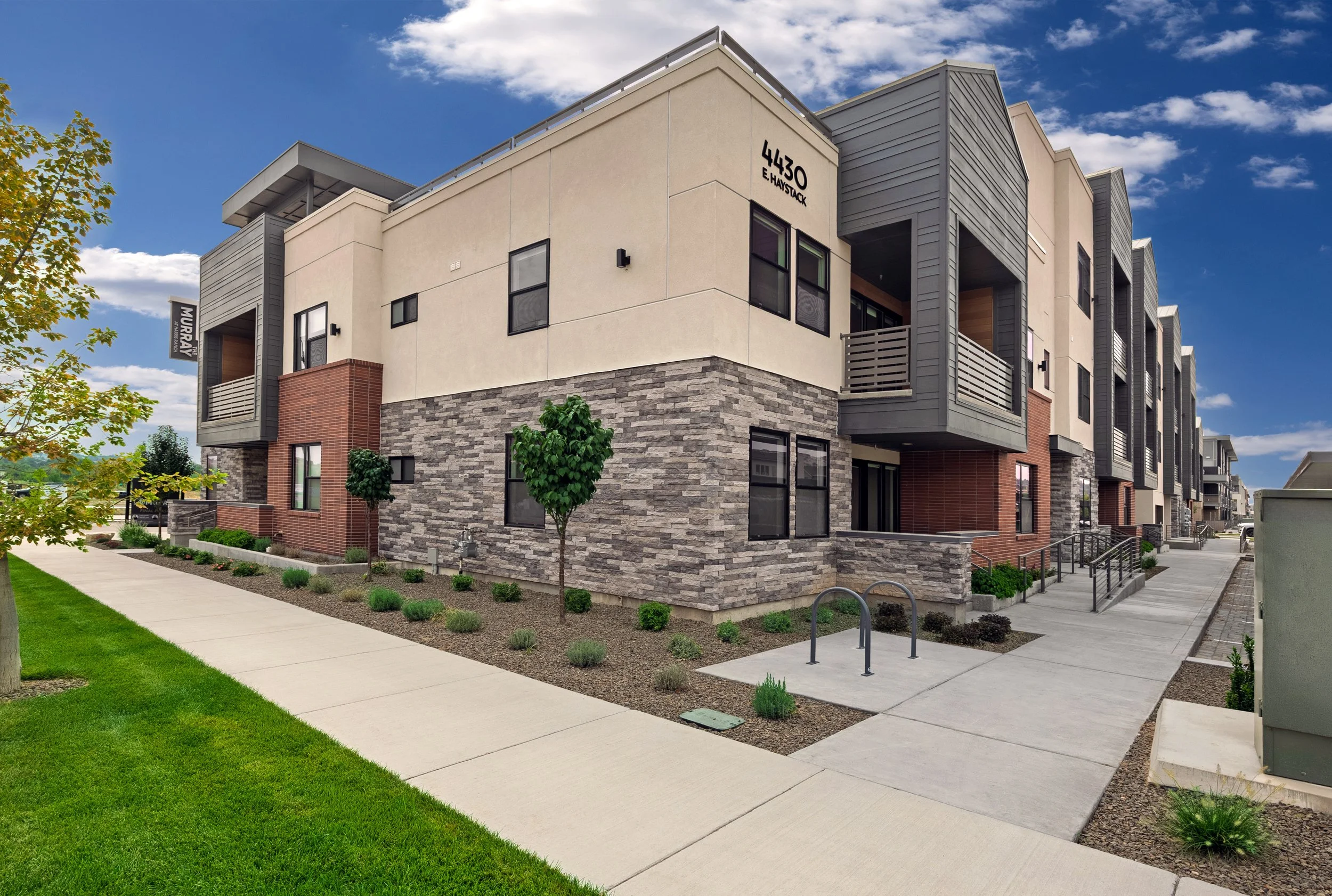 Modern multi-story residential building with mixed exterior finishes including stone, beige stucco, and gray siding, featuring balconies and large windows, situated along a concrete sidewalk with landscaped greenery, small trees, and a blue sky with 