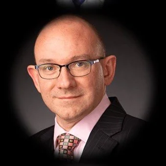 A man with glasses, wearing a dark suit and a patterned tie, posed against a dark background who is a qualified banker and knows a great deal about the retail payments and mass transport industries.