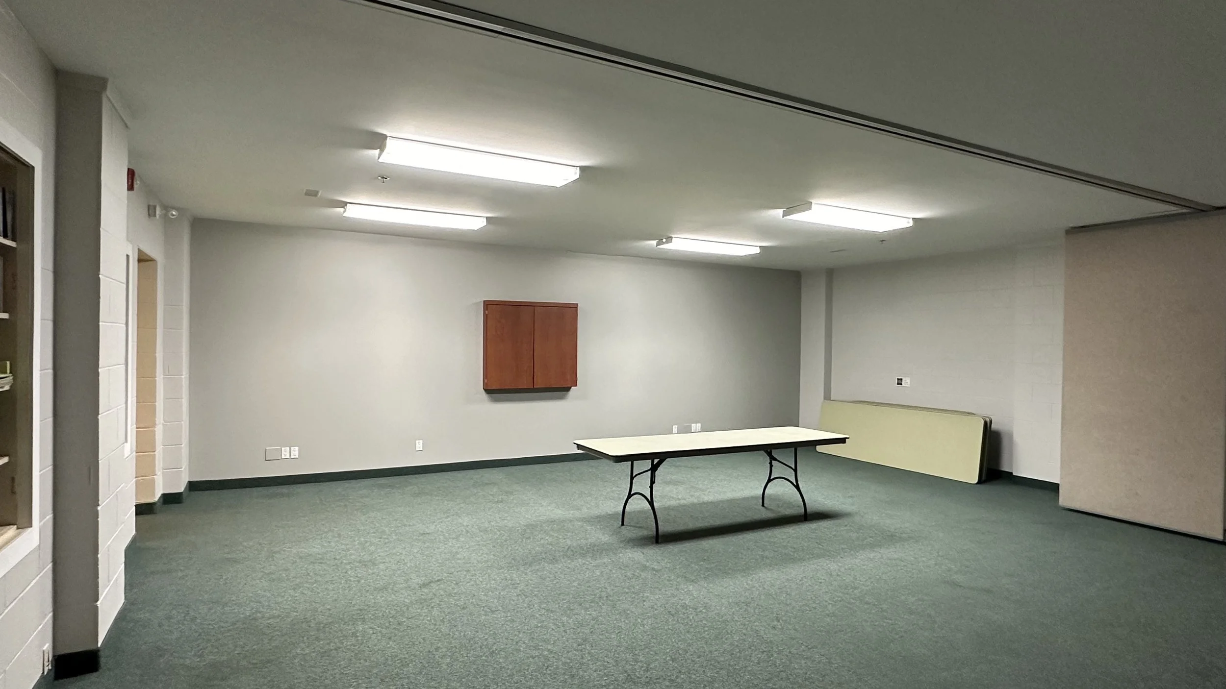 A room with an accordion divider in the centre. A set of blue chairs stacked against the far wall of the room, past the divider. A rectangle table and three blue chairs arranged in the centre of the room. A line of blue chairs arranged against a wall