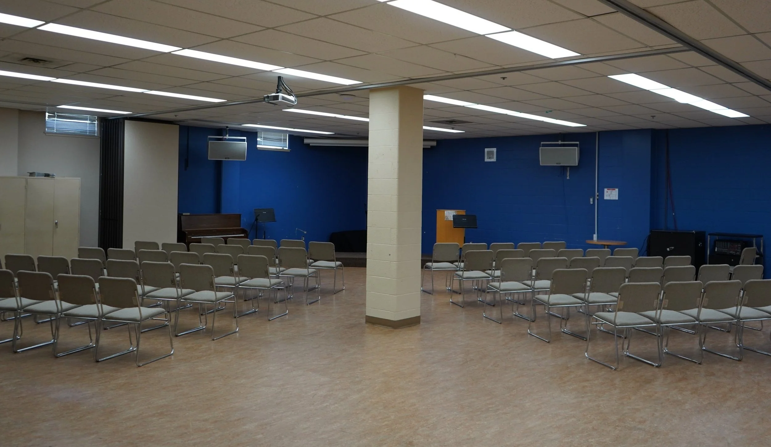 A large open basement space with a blue wall in the far corner, skirting a small riser and a podium. Arrays of grey chairs surround the riser, on either side of a block column.