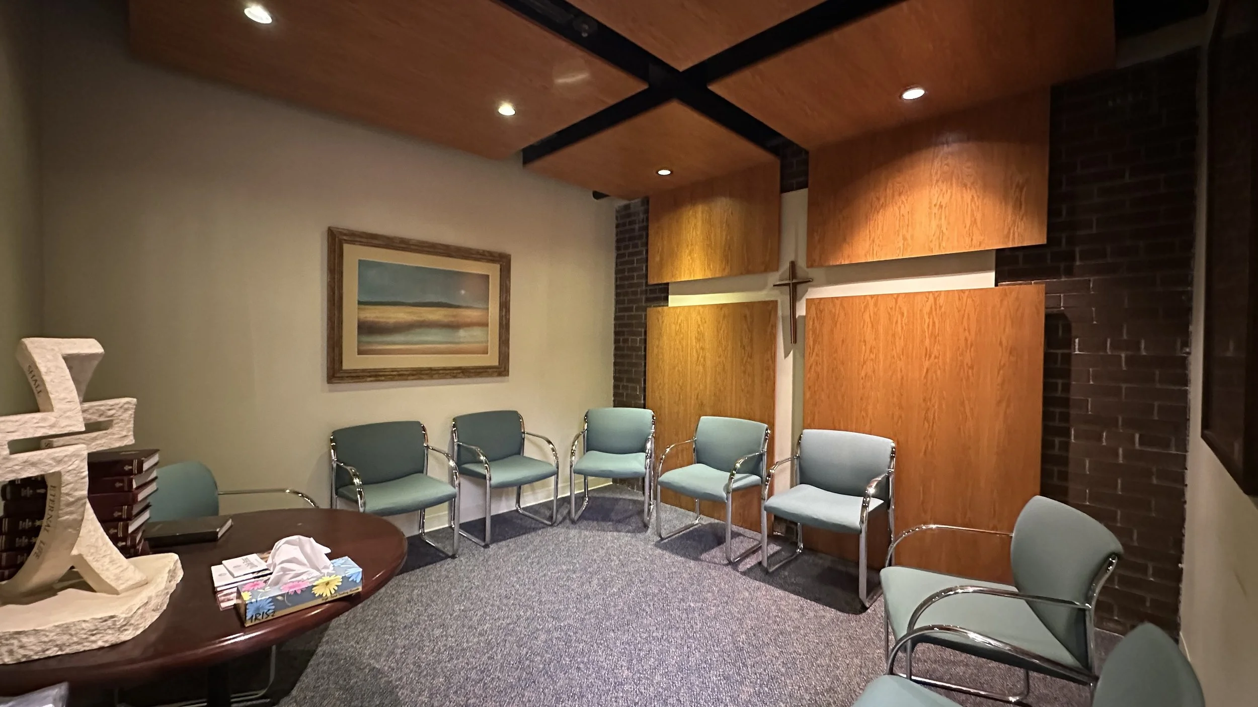 The prayer room with a set of 8 green chairs. A small brown round table. A cross in the centre of four separate wooden panels placed against a dark exposed brick wall.