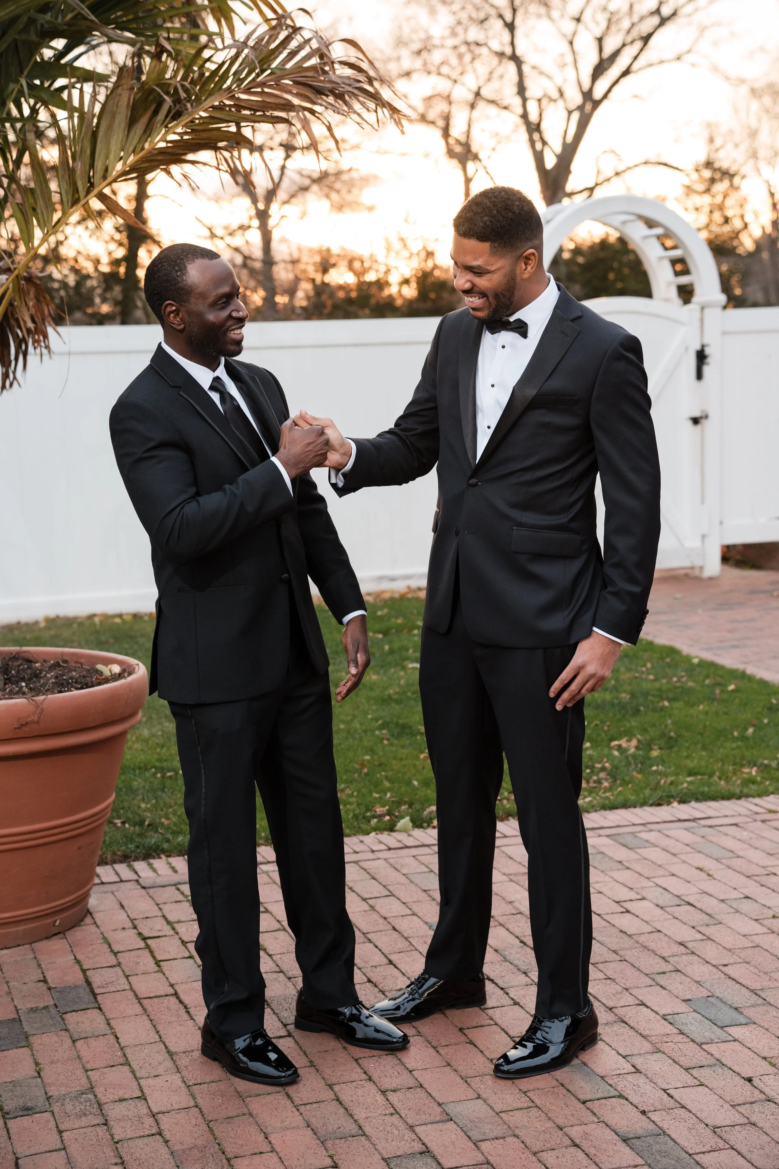 Two men in tuxedos shaking hands and smiling outdoors at sunset.