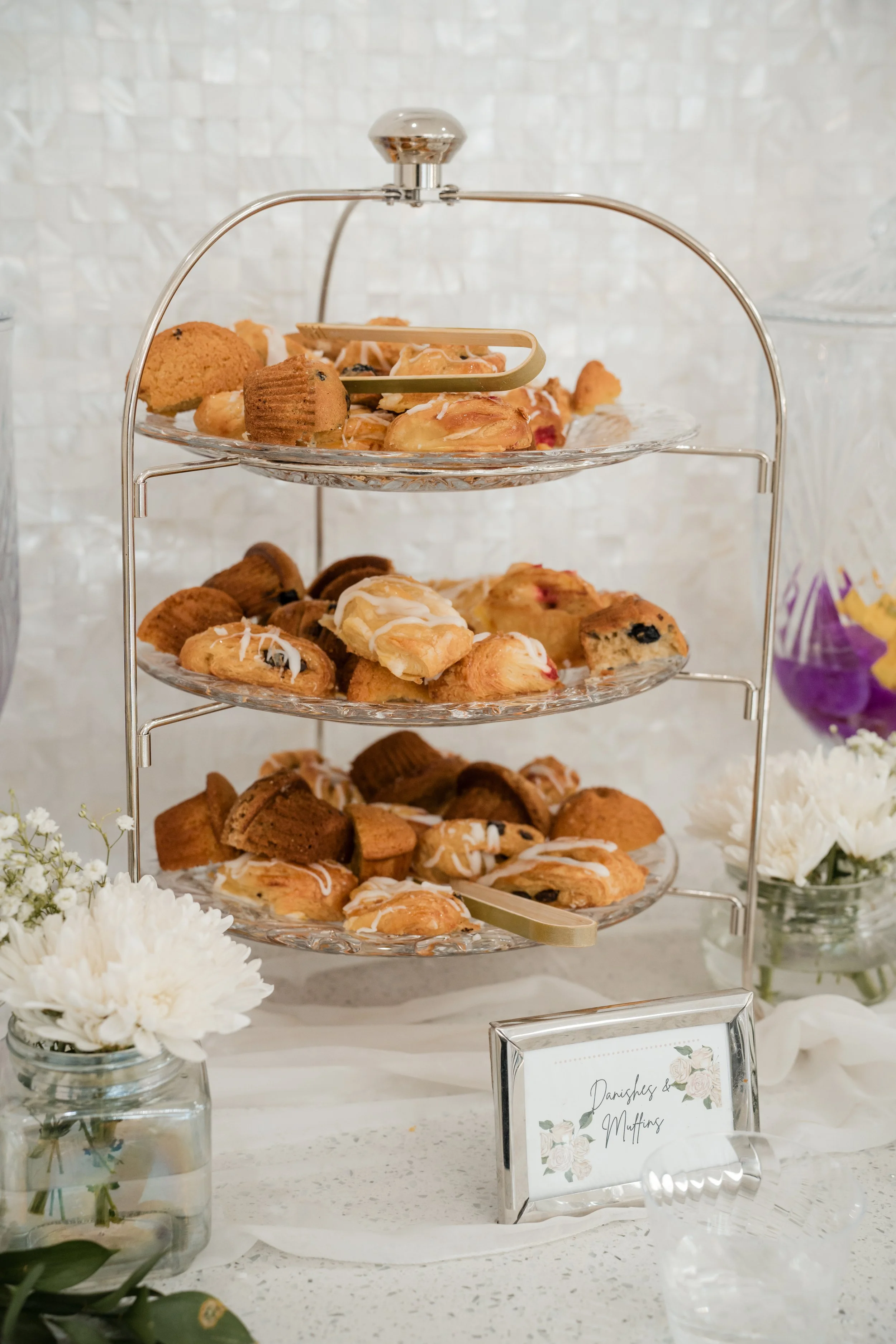 A three-tiered metal and glass tray filled with various pastries, including muffins and cookies, on a white table with white flowers and a sign that reads 'Danishes & Muffins' in a silver frame.