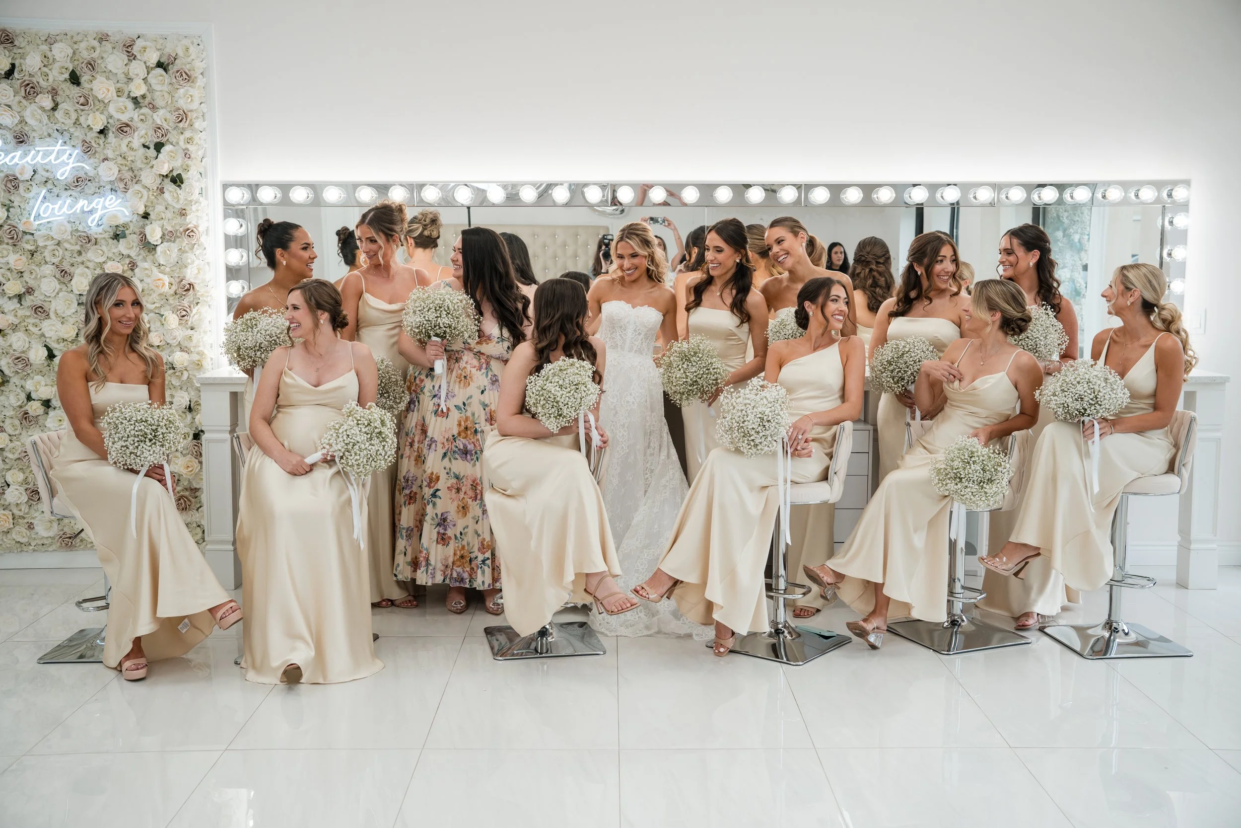 A bride in a white wedding gown surrounded by her bridesmaids in cream-colored dresses, all holding white bouquets, in a makeup and dressing room with a large mirror and floral wall.