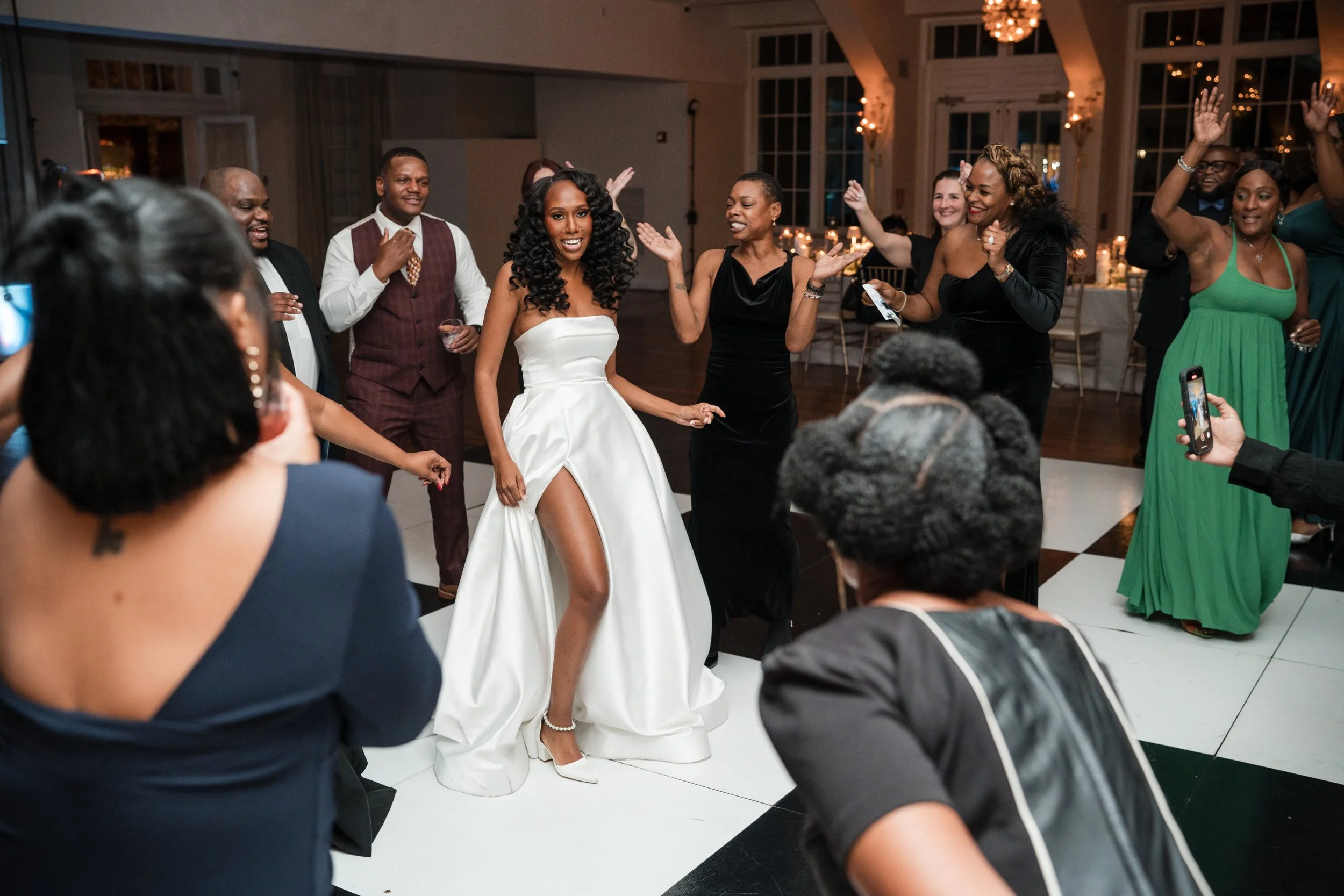 A bride in a strapless white wedding gown with a high slit dancing with guests at her wedding reception. Guests are surrounding her, dancing, smiling, and taking photos in a decorated venue with large windows and warm lighting.