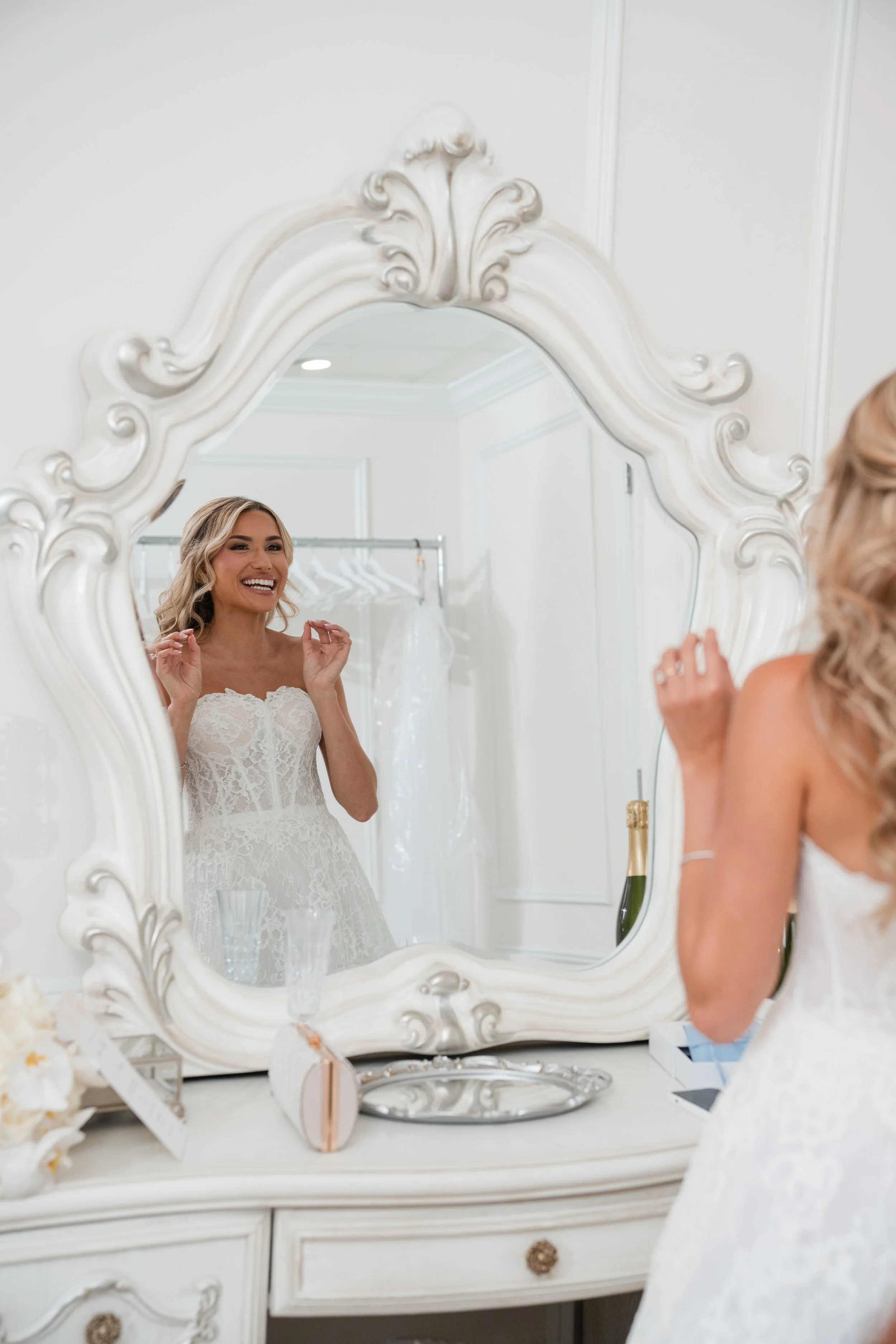 A bride with blonde hair wearing a lace wedding dress, smiling and adjusting her earrings in front of a large ornate mirror.