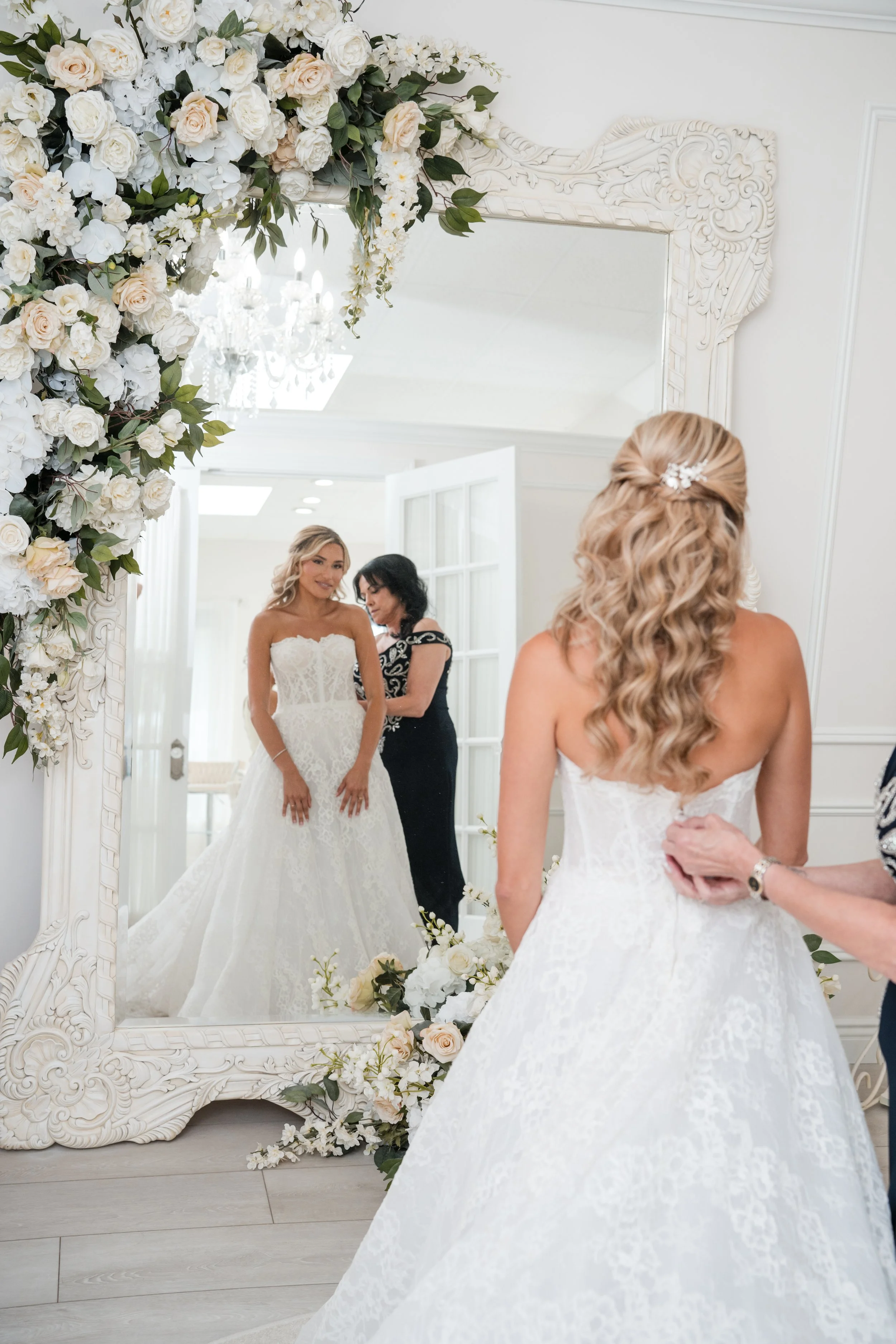 A bride trying on her wedding dress in a fitting room, looking at her reflection in a large ornate mirror decorated with white and blush flowers and greenery, with a woman assisting her.