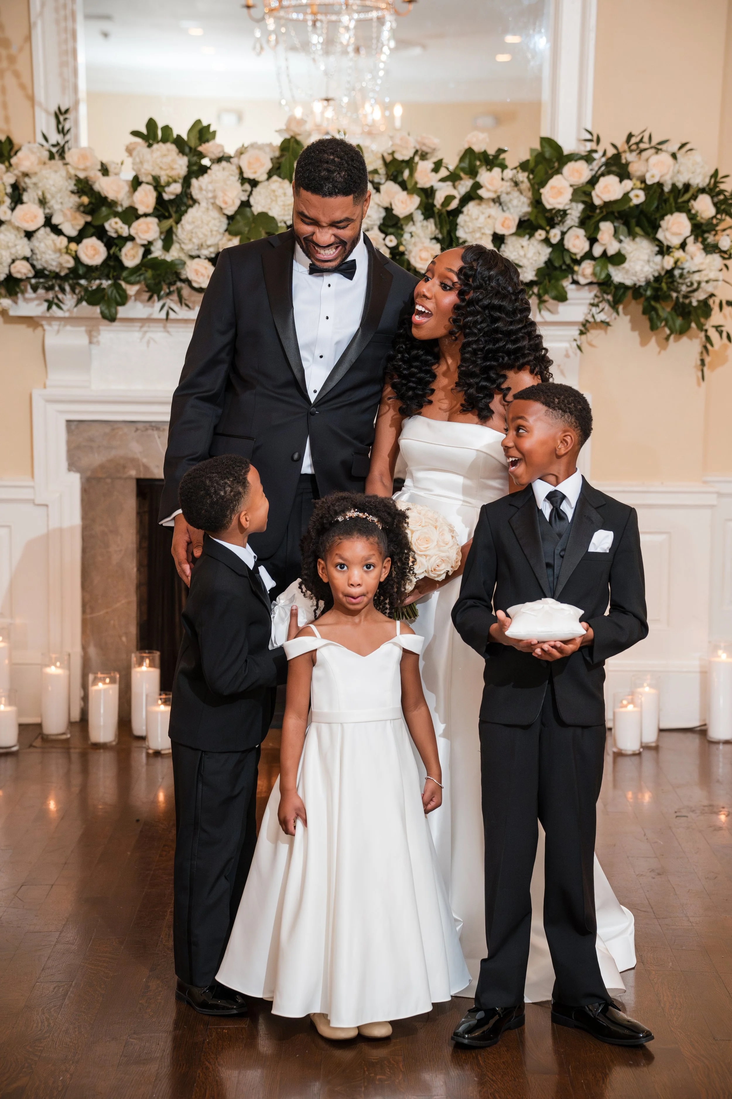 A wedding celebration with a bride, groom, and four children standing in front of a decorated fireplace and floral arrangement, smiling and interacting with each other.