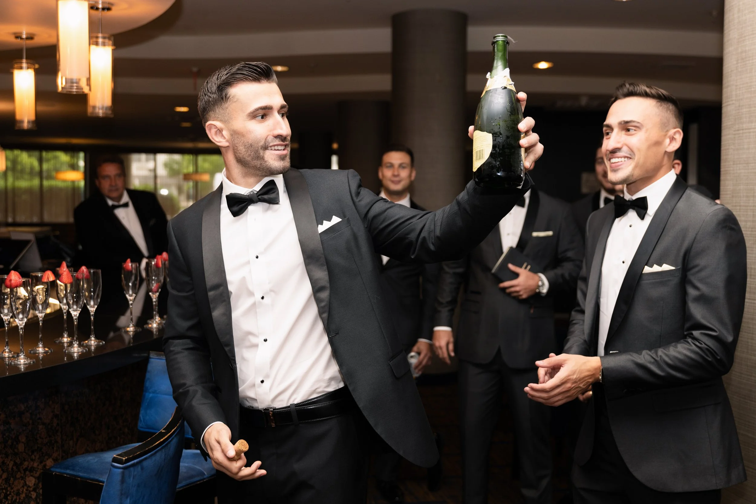 Group of men dressed in tuxedos celebrating at an event, with one man holding a champagne bottle.