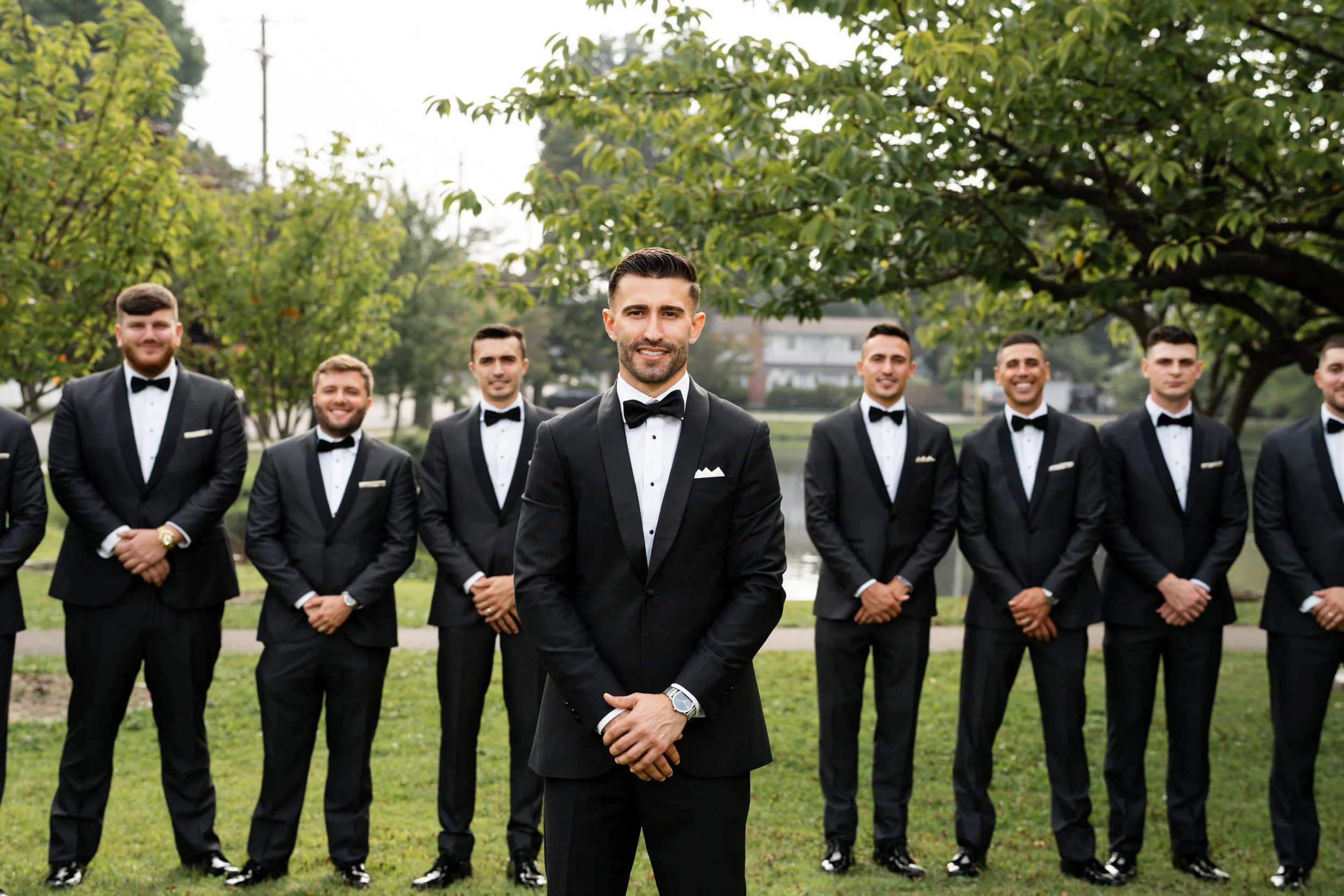 A groom standing in front of his groomsmen outdoors in a park, all dressed in black tuxedos with white shirts and black bow ties, with trees and a lake in the background.