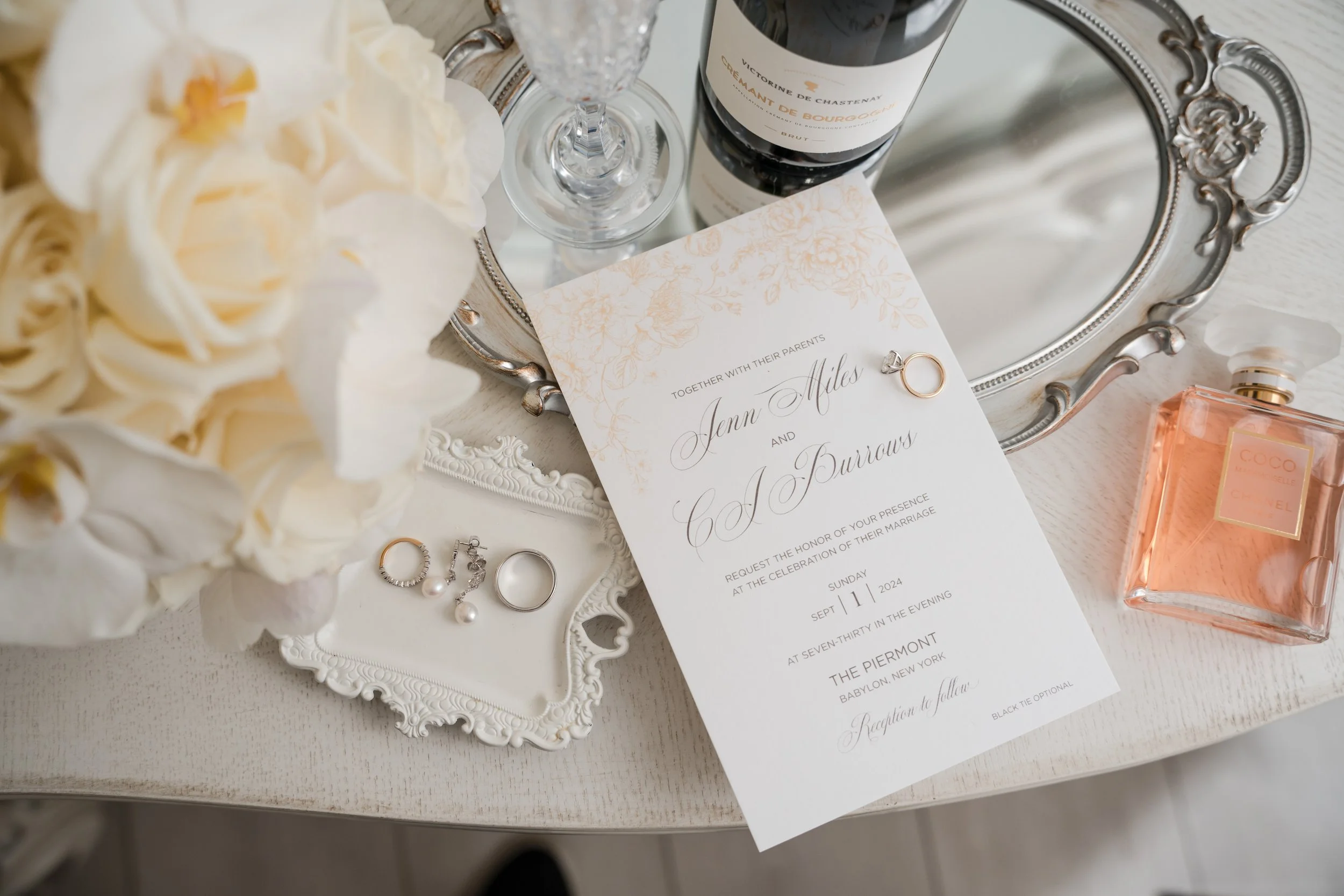 Wedding invitation with rings, jewelry, a bouquet, perfume, wine, and a mirror on a white wooden surface.
