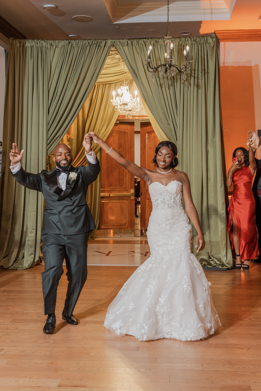 A bride and groom enter their wedding reception. The bride is wearing a strapless white wedding gown, and the groom is dressed in an evergreen tuxedo. They are holding hands and smiling. The background features green curtains, a chandelier.