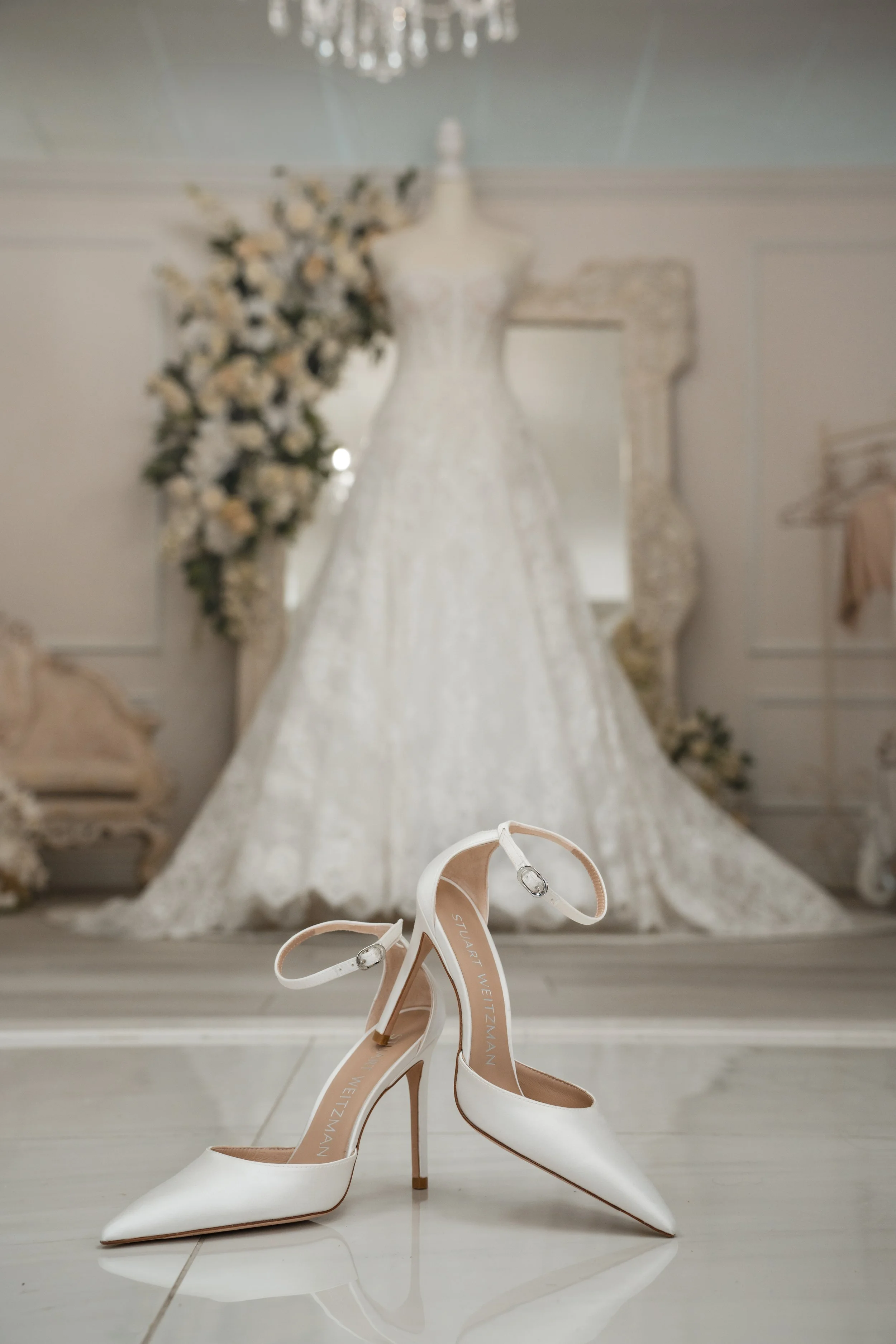 Pair of white high-heeled bridal shoes in front of a wedding dress on a mannequin, with a mirror and floral arrangement in background.