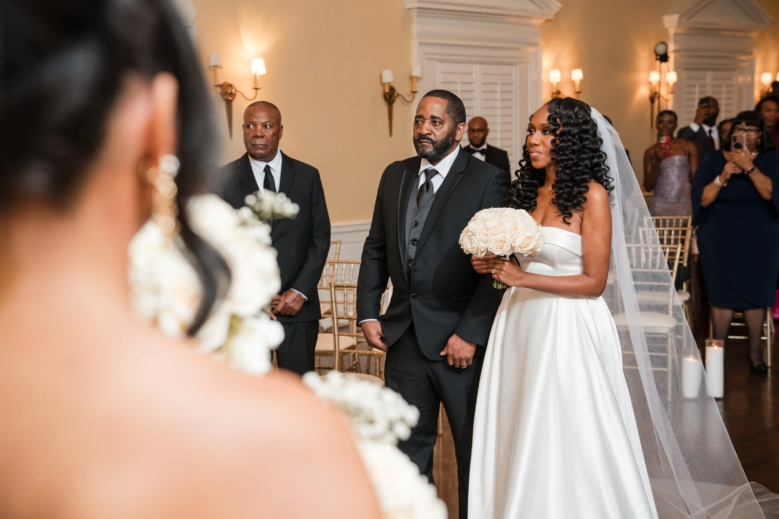 A bride in a white wedding dress holding a bouquet of roses stands next to a man in a black suit during a wedding ceremony. Guests in formal attire are seated and standing in the background.