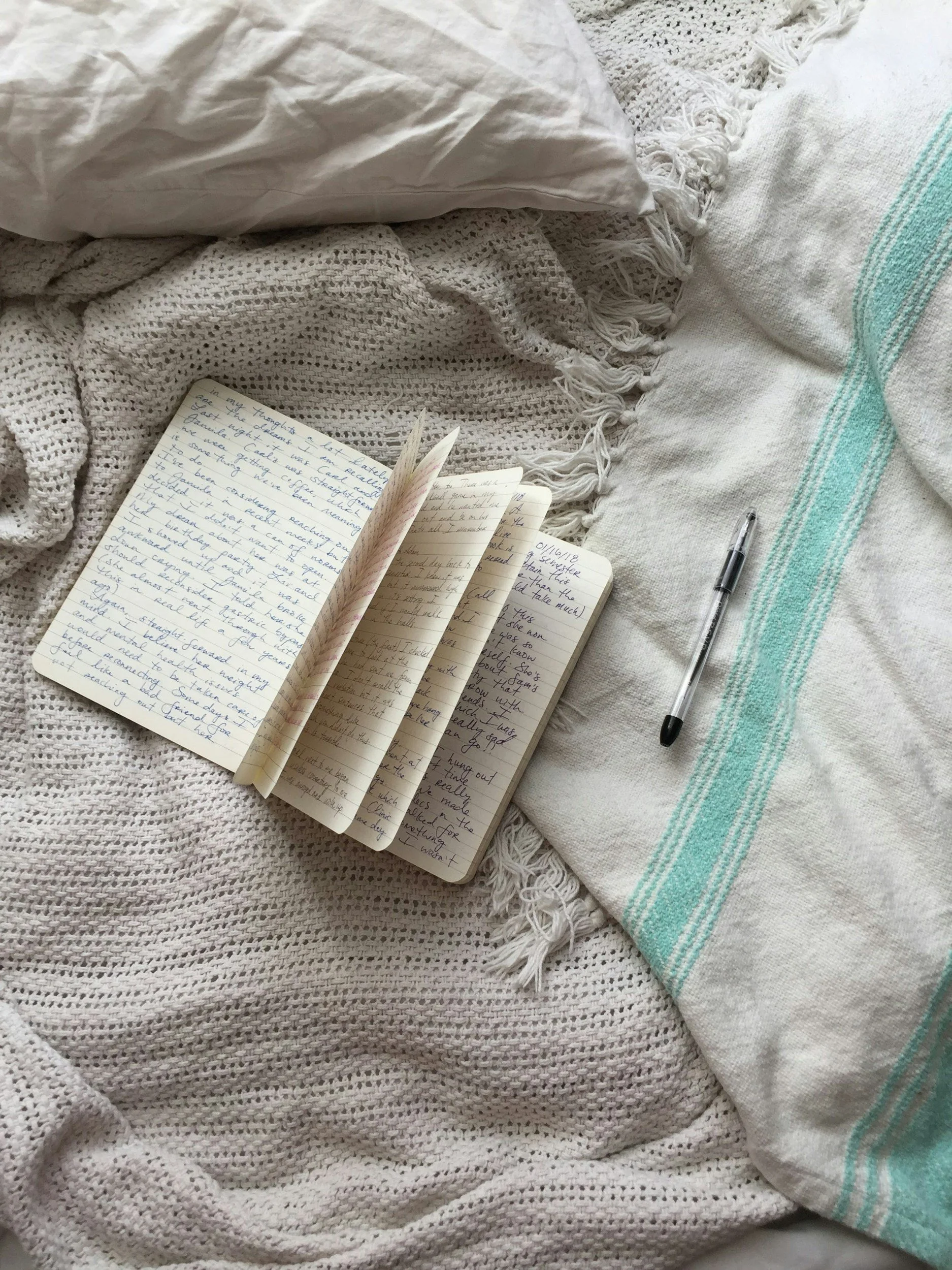 A white pillow, a folded knitted blanket, handwritten notes in a notebook, and a black pen on a textured white blanket with teal stripes.