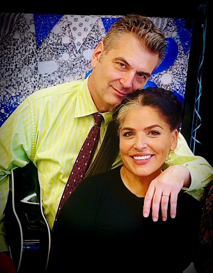 A man with gray hair in a yellow-striped shirt and maroon tie, and a woman with dark hair styled in an updo, smiling. The man is resting his arm on the woman's shoulder. They are in front of a colorful backdrop with blue, white, and gray patterns.