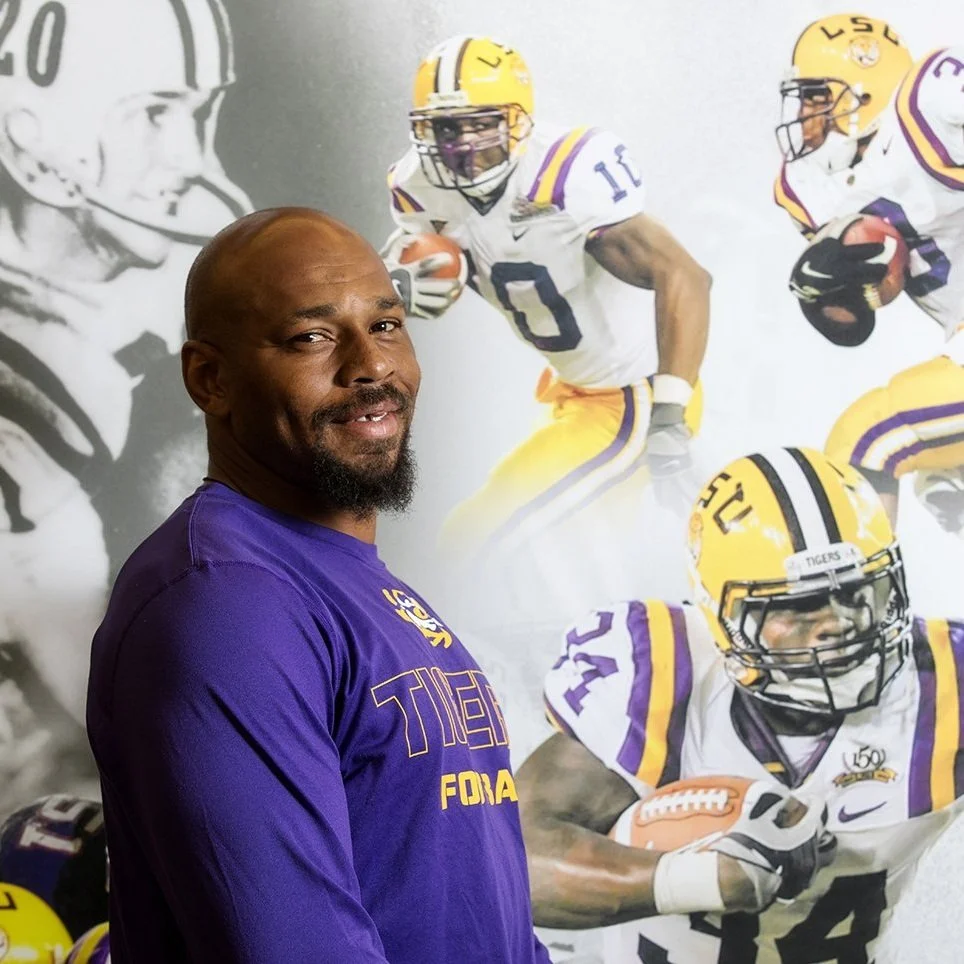 A man in a purple LSU Tigers sweatshirt standing in front of a wall with images of football players in LSU uniforms. The wall features a large grayscale image of a football player and several colored images of players in action.